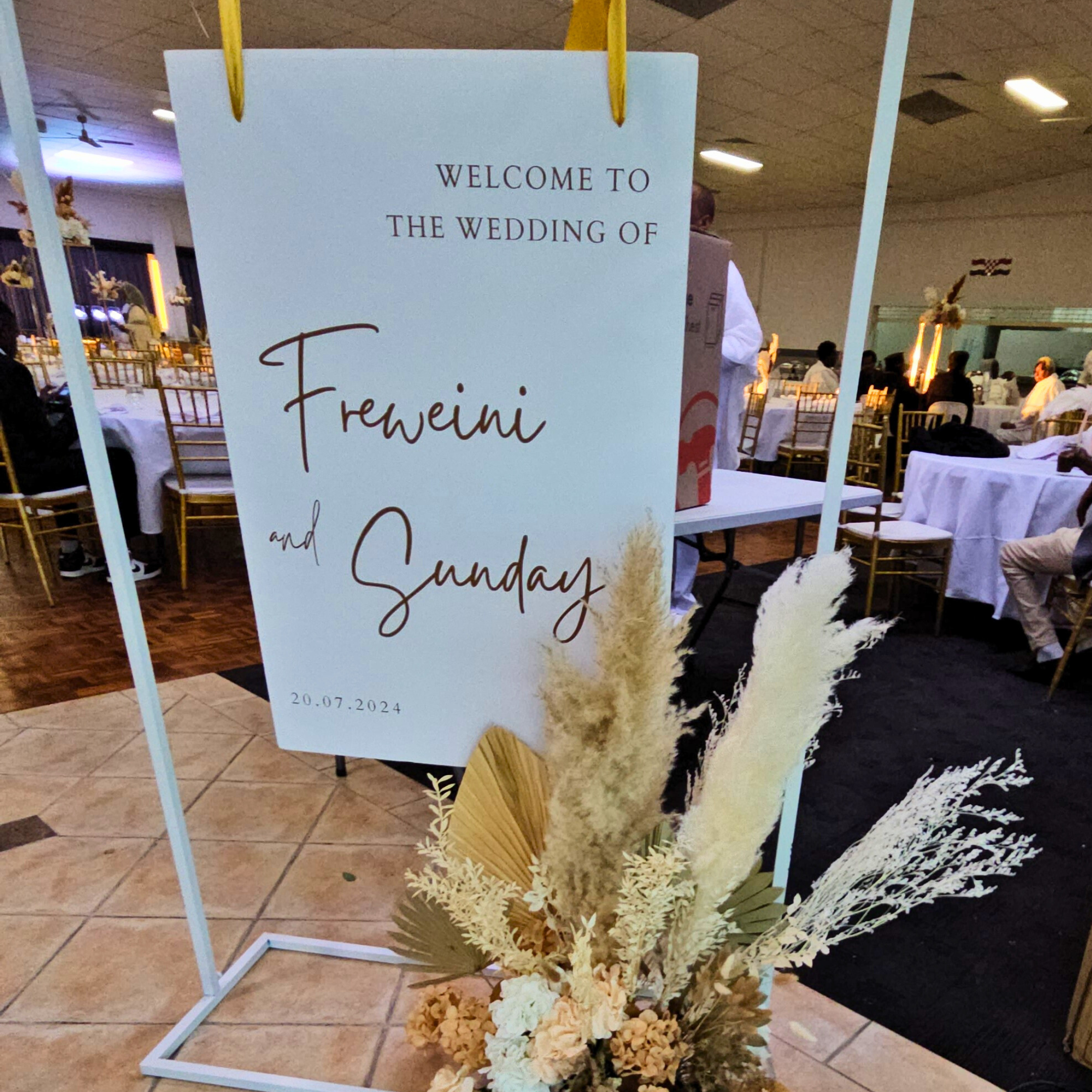 Wedding welcome sign reading ‘WELCOME TO THE WEDDING OF Freweni and Sunday 20.07.2024’ in black script on white, adorned with dried floral arrangements featuring pampas grass and neutral-toned florals. Prop hire, Prop hiring, Prop hire, event stylists Brisbane, brisbane event styling, brisbane event styling, event stylist Brisbane, Brisbane Wedding, Gold Coast Wedding, Floral Styling, Lush & Lavish Events, Brisbane Tablescape, Gold Coast Tablescapes, Brisbane Candle Hire