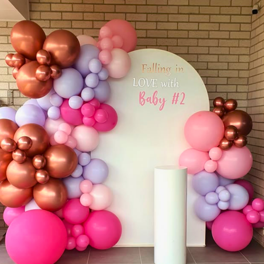 A baby shower backdrop featuring a large white arch with a balloon garland in shades of pink, purple, and rose gold, with the text ' Falling in LOVE with Baby #2' displayed. Backdrop by Lush and Lavish Events a Brisbane and Gold Coast based event styling, event planning and on-the-day coordination business.