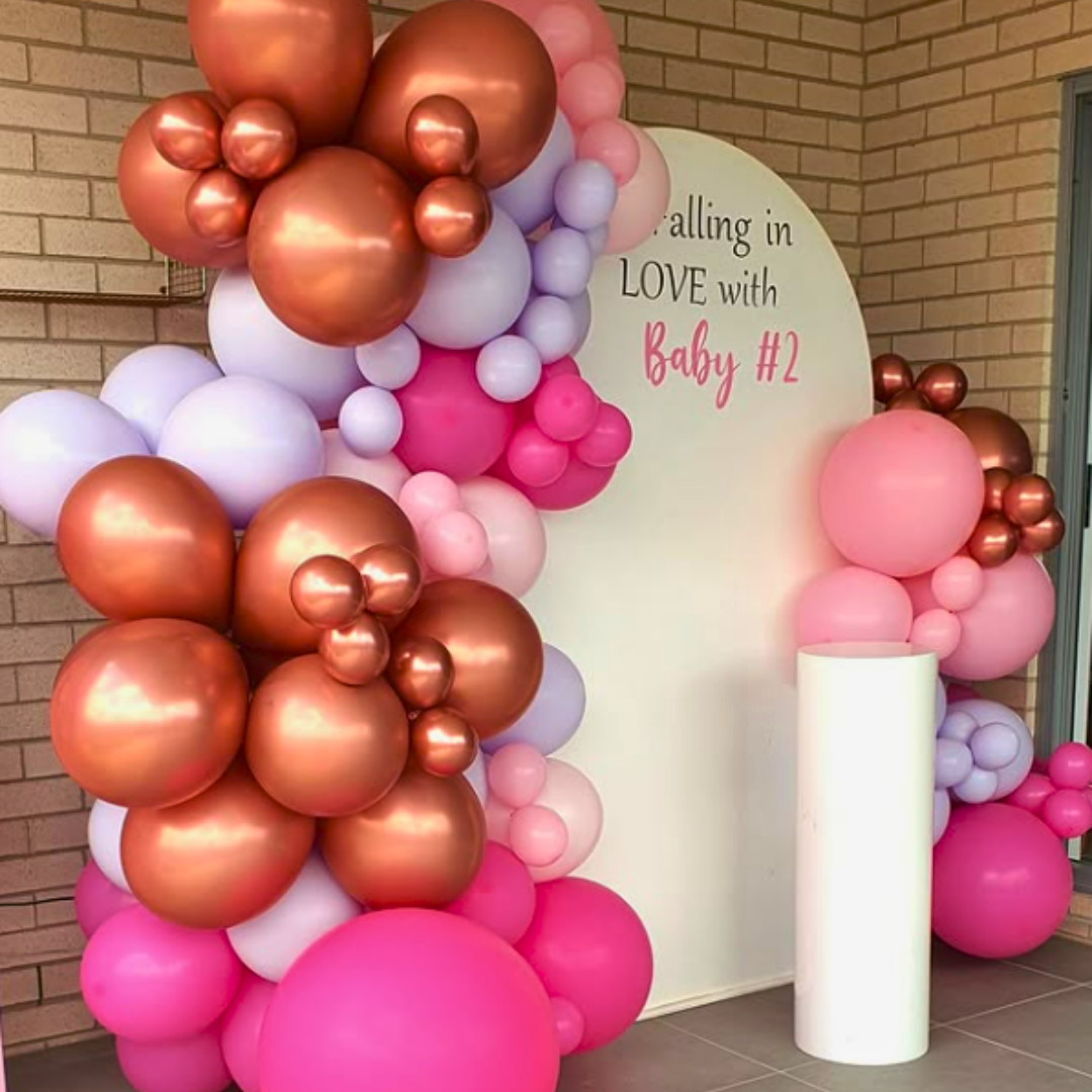 A baby shower backdrop featuring a large white arch with a balloon garland in shades of pink, purple, and rose gold, with the text ' Falling in LOVE with Baby #2' displayed. Backdrop by Lush and Lavish Events a Brisbane and Gold Coast based event styling, event planning and on-the-day coordination business.