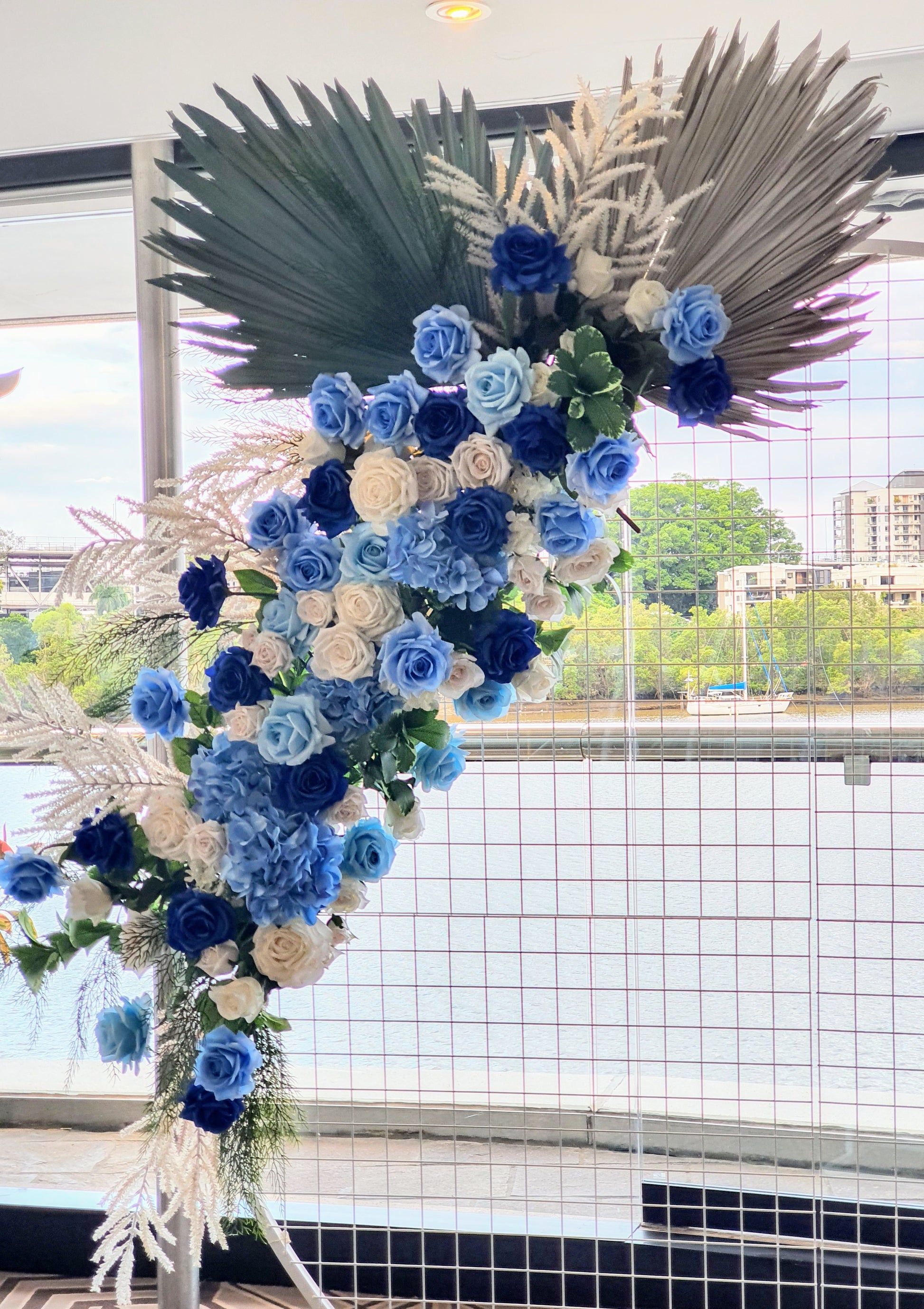 Decorative floral arrangement featuring blue and white roses, green foliage, and white fern-like leaves. The flowers cascade against a wire grid with a river and buildings in the background. Large, dark green palm leaves adorn the top. Prop hire, Prop hiring, Prop hire, event stylists Brisbane, brisbane event styling, brisbane event styling, event stylist Brisbane, Brisbane Wedding, Gold Coast Wedding, Floral Styling, Lush & Lavish Events