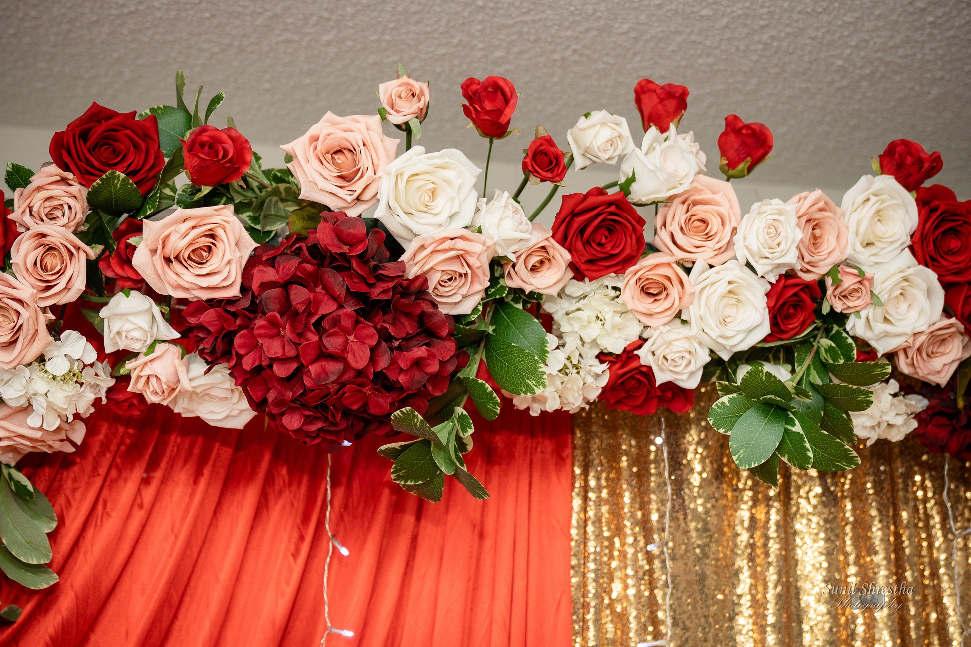 A floral arrangement of roses in shades of red, pink, and white, along with green leaves, set against a backdrop of red and gold fabric. The flowers are arranged in a horizontal line above the fabric. Prop hire, Prop hiring, Prop hire, event stylists Brisbane, brisbane event styling, brisbane event styling, event stylist Brisbane, Brisbane Wedding, Gold Coast Wedding, Floral Styling, Lush & Lavish Events
