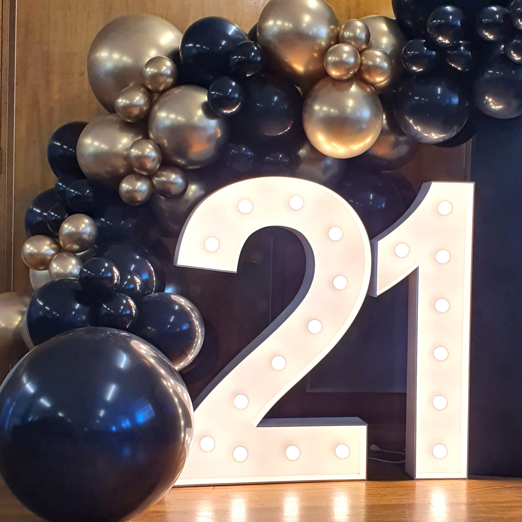 A large illuminated number "21" is displayed in the center of the image, surrounded by a decorative arrangement of black and gold balloons. The balloons are clustered together, creating an arch-like structure above and around the number. The scene appears to be set indoors, likely for a celebration or event marking a 21st birthday or anniversary. Prop hire, Prop hiring, Prop hire, event stylists Brisbane, brisbane event styling, brisbane event styling, event stylist Brisbane, Brisbane Wedding, Gold Coast We