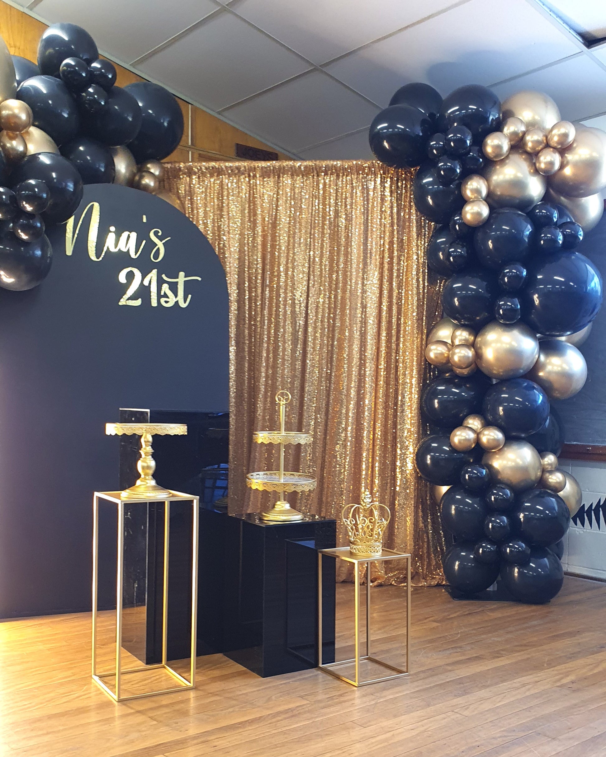 A decorated area for a 21st birthday celebration. The backdrop features a gold sequin curtain with black and gold balloon arrangements on either side. A black arch with the text "Nia's 21st" in gold lettering is on the left. In front of the backdrop, there are three gold stands of varying heights, one with a two-tiered tray and another with a decorative crown. Prop hire, Prop hiring, Prop hire, event stylists Brisbane, brisbane event styling, brisbane event styling, event stylist Brisbane, Brisbane Wedding,