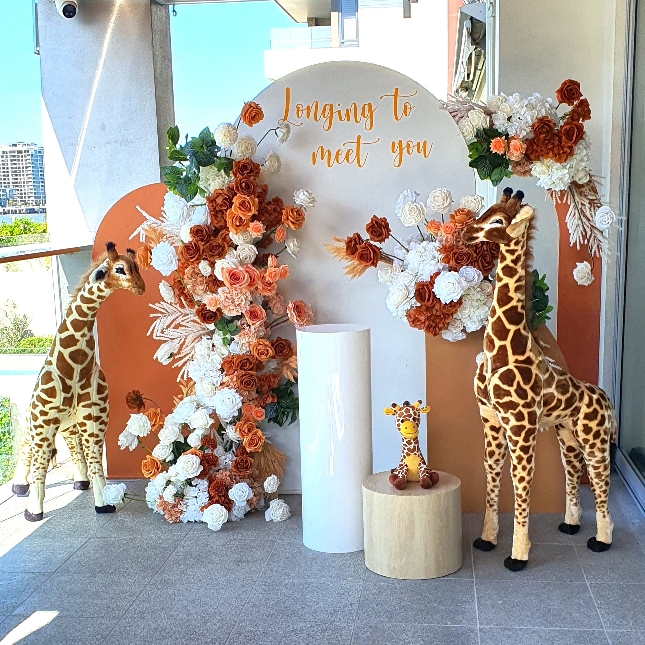 Two large giraffe statues and one small giraffe statue on a wooden stool. The backdrop includes a white and orange arch with orange and white flowers, with the text “longing to meet you.” Two large giraffe statues and one small giraffe statue on a wooden stool. The backdrop includes a white and orange arch with orange and white flowers, with the text “longing to meet you.”