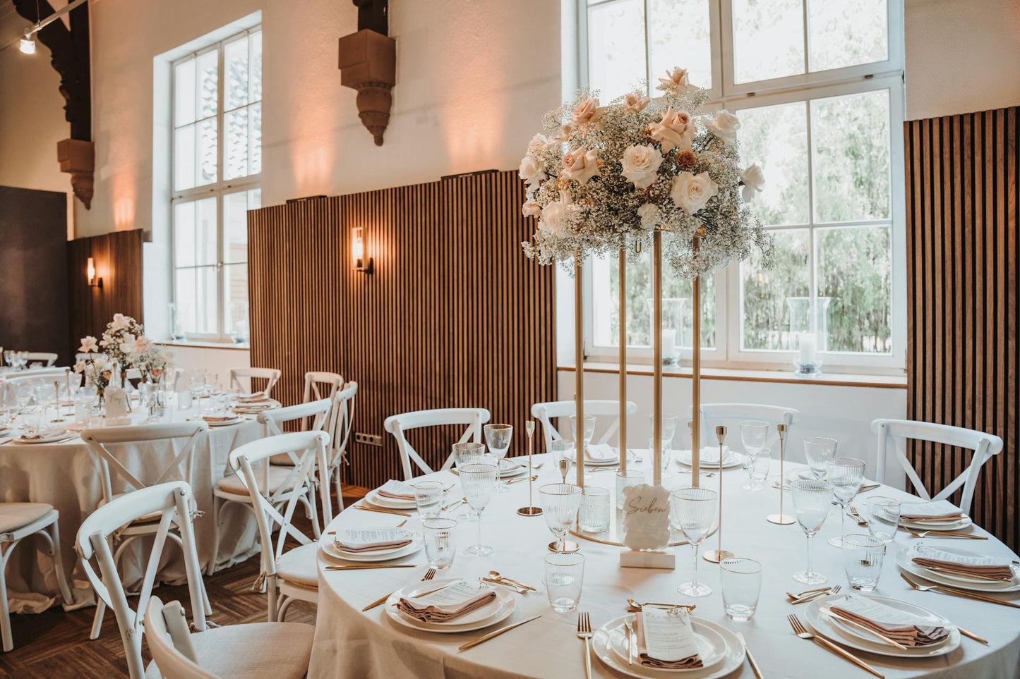 Event space with round tables featuring white tablecloths, elegant table settings, and tall floral centerpieces of white and blush flowers. Large windows let in natural light, with wooden panels and sconces on the walls. Prop hire, Prop hiring, Prop hire, event stylists Brisbane, brisbane event styling, brisbane event styling, event stylist Brisbane, Brisbane Wedding, Gold Coast Wedding, Floral Styling, Lush & Lavish Events