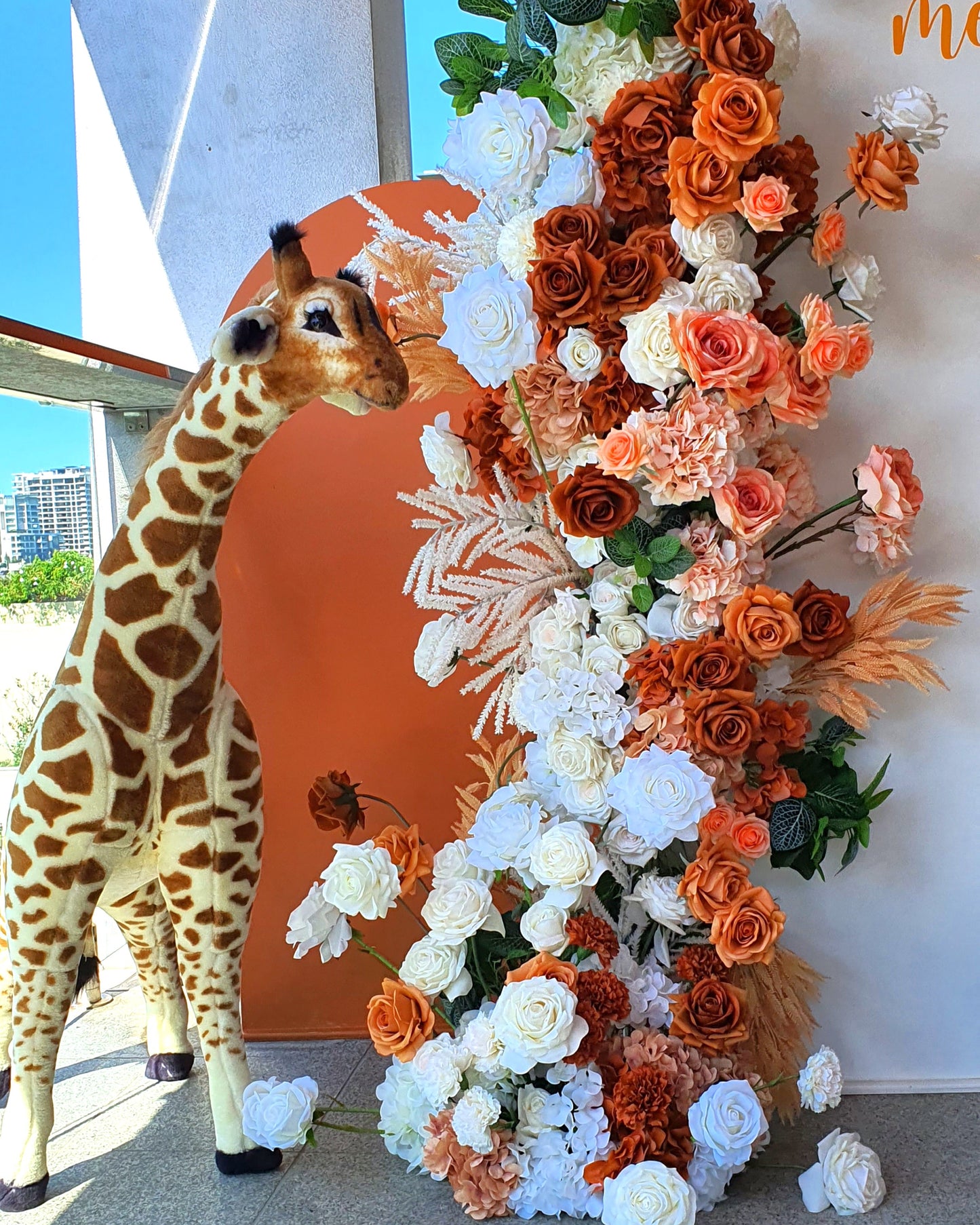 Two large giraffe statues and one small giraffe statue on a wooden stool. The backdrop includes a white and orange arch with orange and white flowers, with the text “longing to meet you.” Prop hire, Prop hiring, Prop hire, event stylists Brisbane, brisbane event styling, brisbane event styling, event stylist Brisbane, Brisbane Wedding, Gold Coast Wedding, Floral Styling, Lush & Lavish Events, Brisbane Tablescape, Gold Coast Tablescapes, Brisbane Candle Hire