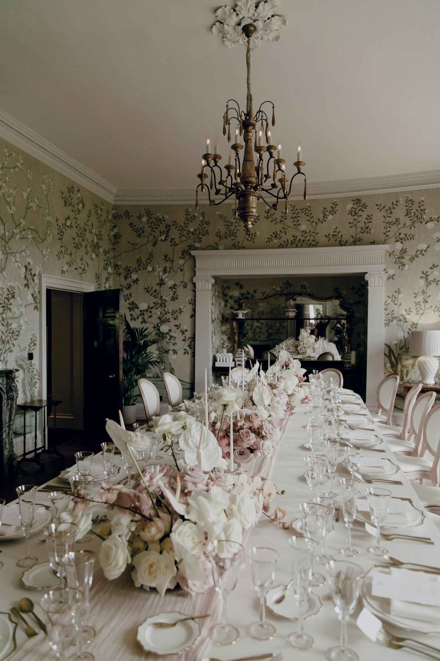 A long dining table is set with white tablecloths and silver cutlery. A stunning floral centerpiece of white and pink flowers runs down the center of the table, and a grand chandelier hangs from the ceiling. Prop hire, Prop hiring, Prop hire, event stylists Brisbane, brisbane event styling, brisbane event styling, event stylist Brisbane, Brisbane Wedding, Gold Coast Wedding, Floral Styling, Lush & Lavish Events, Brisbane Tablescape, Gold Coast Tablescapes, Brisbane Candle Hire