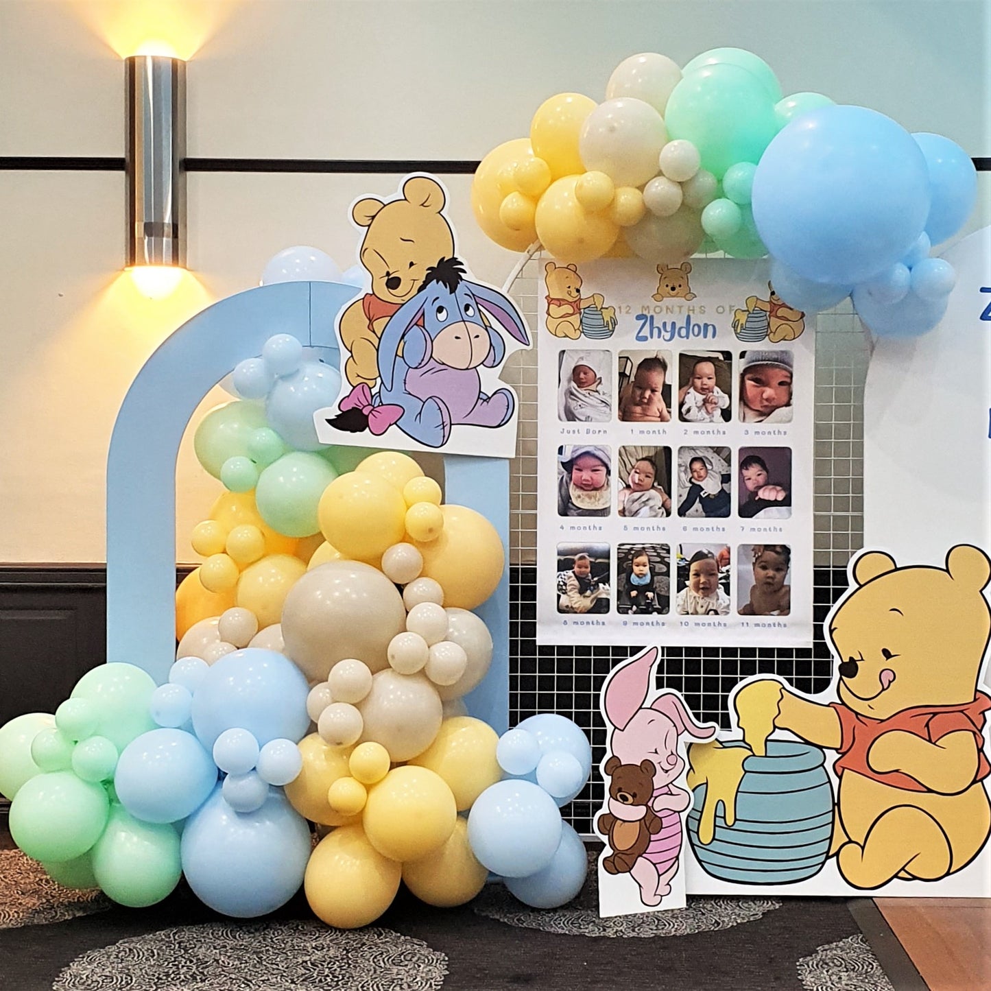 A Winnie the Pooh themed birthday party backdrop features a balloon arch in blue, yellow, and white. Cutouts of Winnie the Pooh characters, and a photo collage of the birthday child. Prop hire, Prop hiring, Prop hire, event stylists Brisbane, brisbane event styling, brisbane event styling, event stylist Brisbane, Brisbane Wedding, Gold Coast Wedding, Floral Styling, Lush & Lavish Events, Brisbane Tablescape, Gold Coast Tablescapes, Brisbane Candle Hire