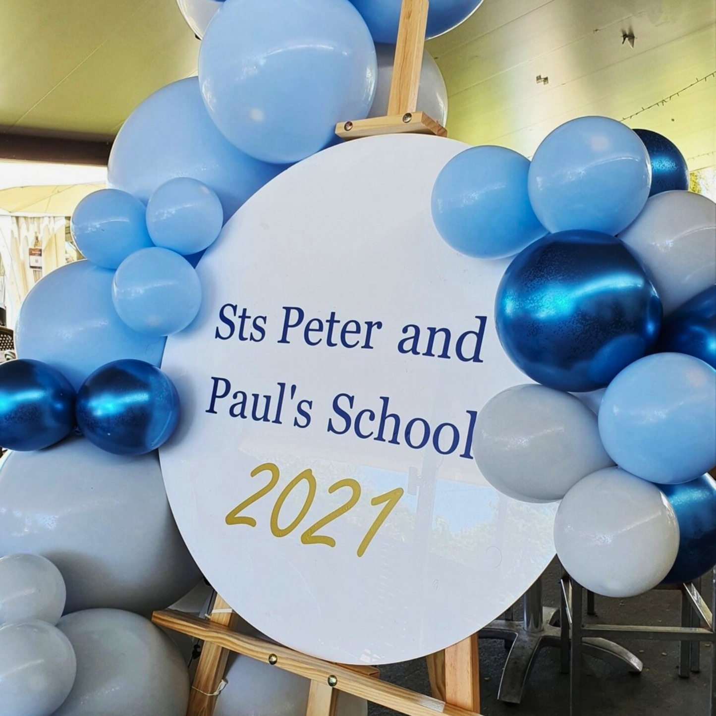 Cluster of blue and white balloons around a sign reading ‘Sts Peter and Paul’s School 2021’ on an easel. Outdoor setting with chairs and tables under a covered area, suggesting a school event or ceremony. Prop hire, Prop hiring, Prop hire, event stylists Brisbane, brisbane event styling, brisbane event styling, event stylist Brisbane, Brisbane Wedding, Gold Coast Wedding, Floral Styling, Lush & Lavish Events, Brisbane Corporate Styling, Gold Coast Corporate Styling, Brisbane Brand Activation