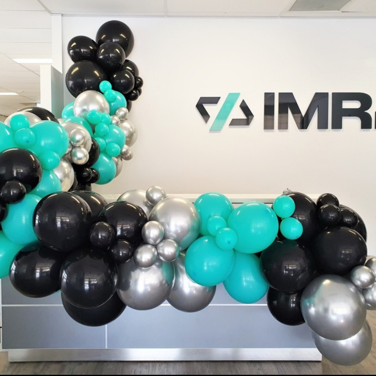 Indoor setting with a wall-mounted ‘IMR’ logo flanked by black, teal, and metallic silver balloons, arranged organically, suggesting a professional yet festive event. Prop hire, Prop hiring, Prop hire, event stylists Brisbane, brisbane event styling, brisbane event styling, event stylist Brisbane, Brisbane Wedding, Gold Coast Wedding, Floral Styling, Lush & Lavish Events, Brisbane Corporate Styling, Gold Coast Corporate Styling, Brisbane Brand Activation