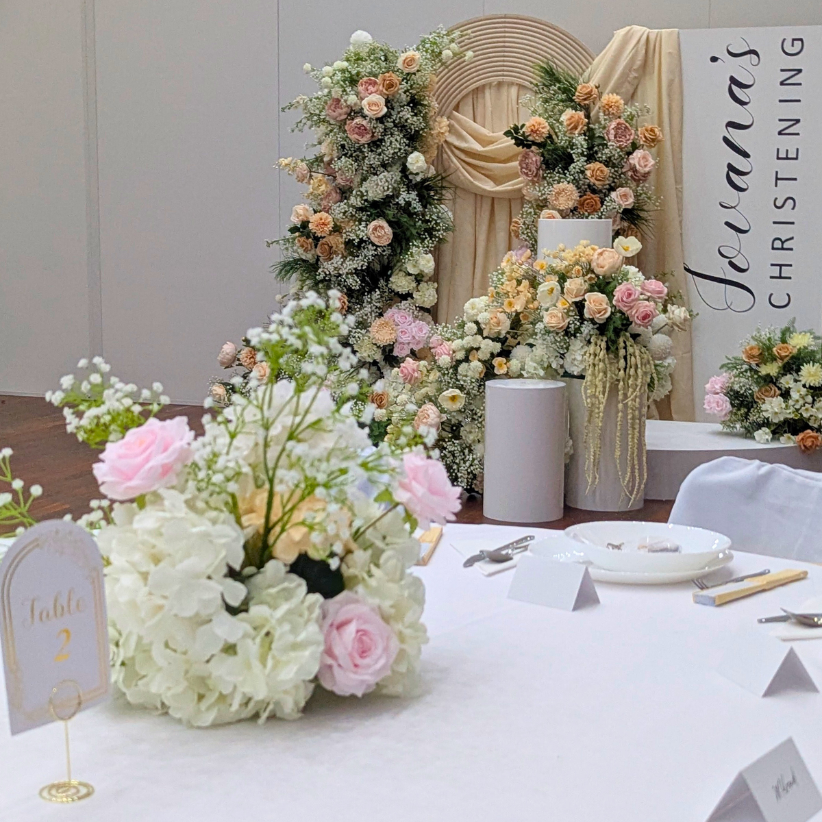 Decorative floral arrangements on a table with a 'Jovana's Christening' sign in the background. christening backdrop hire, baptism backdrop hire, floral backdrop hire Brisbane, floral backdrop hire Gold Coast, christening styling Brisbane, baptism styling Gold Coast, white floral backdrop, nude pink floral backdrop, luxury christening decor, elegant baptism decorations, church christening backdrop, baby christening decorations, event backdrop hire Brisbane, event backdrop hire Gold Coast, premium floral wa
