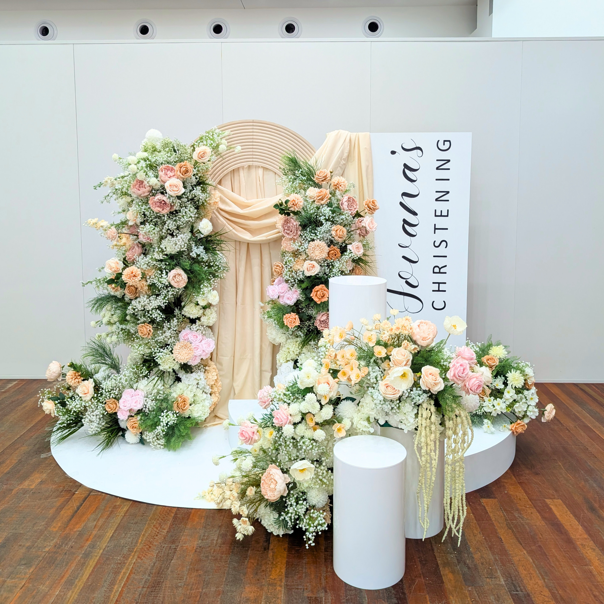 Decorative floral arrangement with a 'Jocelina's Christening' sign on a wooden floor. christening backdrop hire, baptism backdrop hire, floral backdrop hire Brisbane, floral backdrop hire Gold Coast, christening styling Brisbane, baptism styling Gold Coast, white floral backdrop, nude pink floral backdrop, luxury christening decor, elegant baptism decorations, church christening backdrop, baby christening decorations, event backdrop hire Brisbane, event backdrop hire Gold Coast, premium floral wall hire, ch