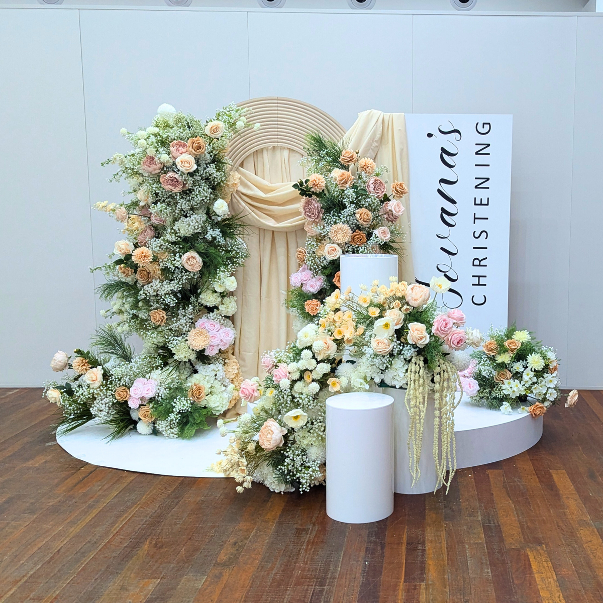 Decorative floral arrangement with a sign indicating a christening celebration on a wooden floor. christening backdrop hire, baptism backdrop hire, floral backdrop hire Brisbane, floral backdrop hire Gold Coast, christening styling Brisbane, baptism styling Gold Coast, white floral backdrop, nude pink floral backdrop, luxury christening decor, elegant baptism decorations, church christening backdrop, baby christening decorations, event backdrop hire Brisbane, event backdrop hire Gold Coast, premium floral w