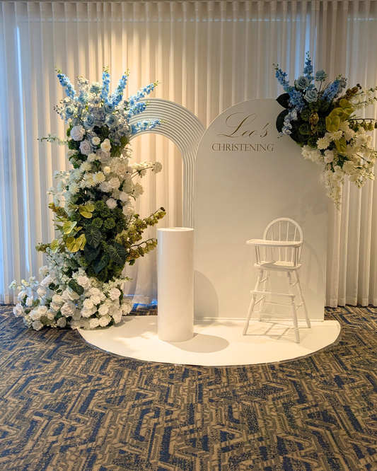 A custom floral Christening backdrop setup featuring a white arched background with 'Leo's CHRISTENING' printed on it, flanked by large floral arrangements in white, blue, and green, with a white pedestal and a white wooden high chair on a patterned carpet. Styled by Brisbane based event stylists, event planners and event coordinators Lush and Lavish Events.
