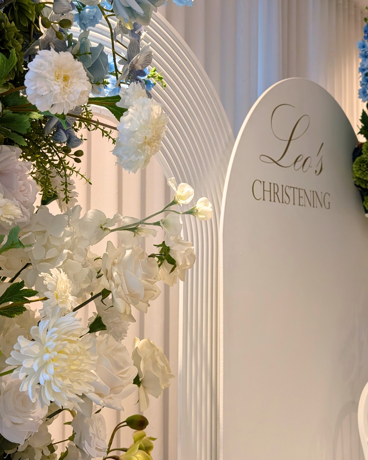 A close-up of a white arched backdrop with the words 'Leo's Christening' in gold text, decorated with large white and pale blue flowers and greenery on the left. Styled by Brisbane based event stylists, event planners and event coordinators Lush and Lavish Events.
