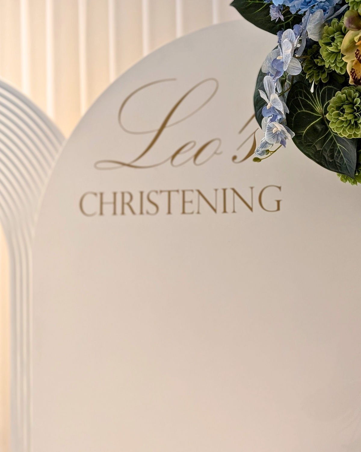 A close-up of a white-colored arch backdrop with elegant gold foil lettering that reads 'Leo's CHRISTENING', decorated with a floral arrangement of blue florals and green foliag. Styled by Brisbane based event stylists, event planners and event coordinators Lush and Lavish Events.