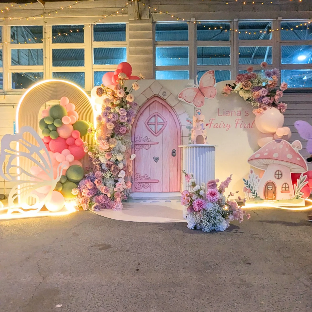Decorative setup with fairy-themed elements including a pink door, balloons, and flowers. fairy party backdrop, enchanted birthday backdrop, fairy first birthday, whimsical party decor, Brisbane party hire, floral fairy arch, magical event styling, fairyland backdrop hire, pastel birthday theme, enchanted garden party decorations