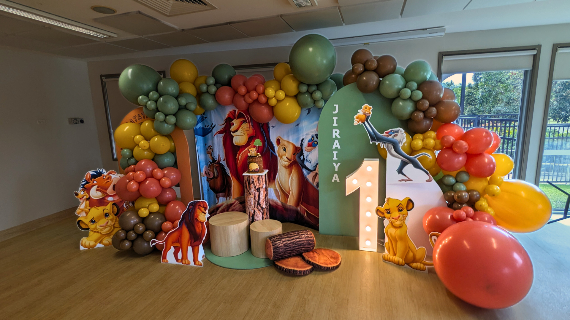 Colorful Lion King themed party backdrop featuring life-sized cutouts of Simba, Nala, and other characters, with ‘JIRAIYA’ spelled out in large letters amidst a vibrant balloon arch. The setting is indoors with natural light coming through windows Lush and Lavish Events