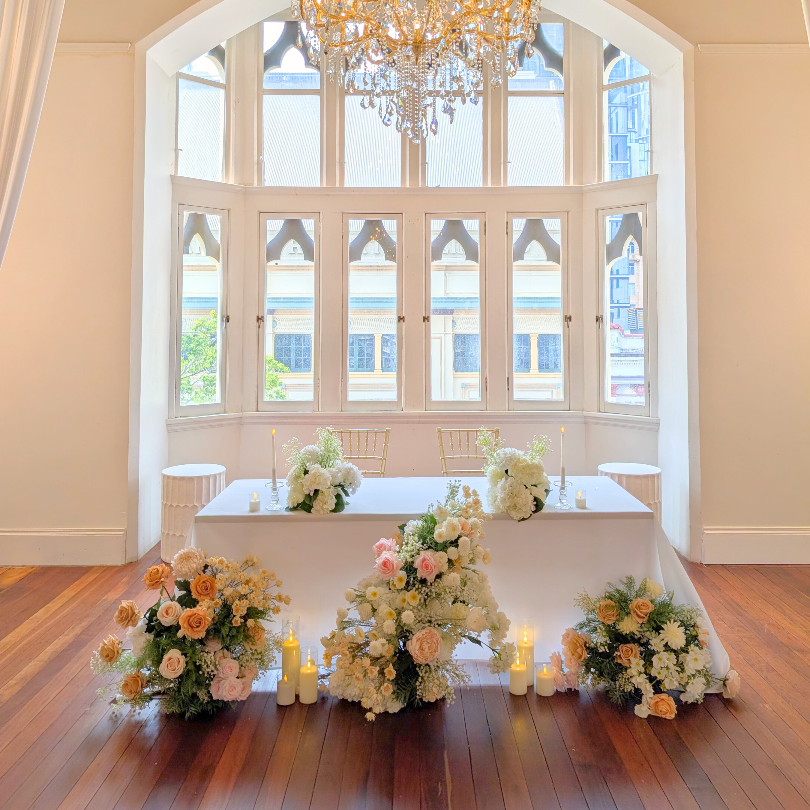 Bridal table with floral stylingand candles in reception room with large windows and a chandelier. bridal table styling brisbane, wedding bridal table decor ideas, bridal table styling inspiration, bridal table floral styling, wedding sweetheart table styling, bridal table candle styling, bridal table backdrop styling, bridal table flowers and candles, bridal table decor australia, wedding head table styling ideas, wedding reception styling brisbane, reception table styling ideas, wedding reception decor.
