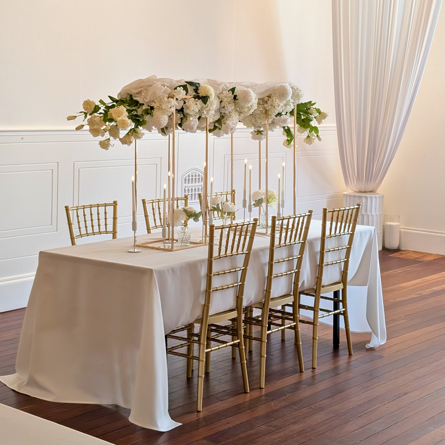 Elegant table setting with white floral centerpiece, candles, and gold chairs in a formal room. brisbane wedding guest table styling, brisbane wedding centrepieces, taper candle wedding styling brisbane, candelabra wedding centrepieces brisbane, bud vase wedding styling brisbane, tall floral plinth centrepieces brisbane, luxury guest table styling brisbane, wedding table centrepiece hire brisbane, modern wedding table styling brisbane, elegant reception table styling brisbane, wedding candle centrepieces.