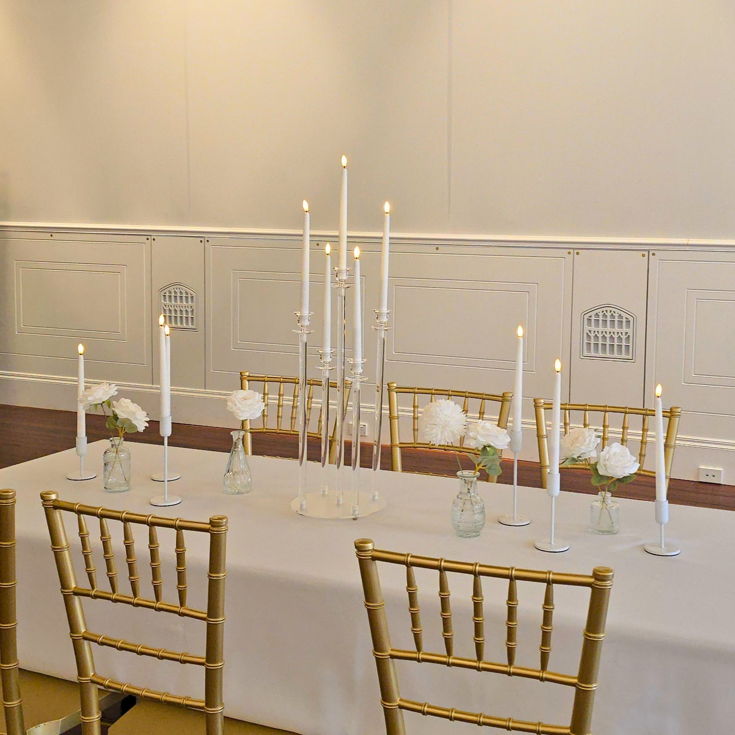 Decorated table with white tablecloth, gold chairs, taper candles, and floral arrangements in a reception room. brisbane wedding guest table styling, brisbane wedding centrepieces, taper candle wedding styling brisbane, candelabra wedding centrepieces brisbane, bud vase wedding styling brisbane, tall floral plinth centrepieces brisbane, luxury guest table styling brisbane, wedding table centrepiece hire brisbane, modern wedding table styling brisbane, elegant reception table styling brisbane.