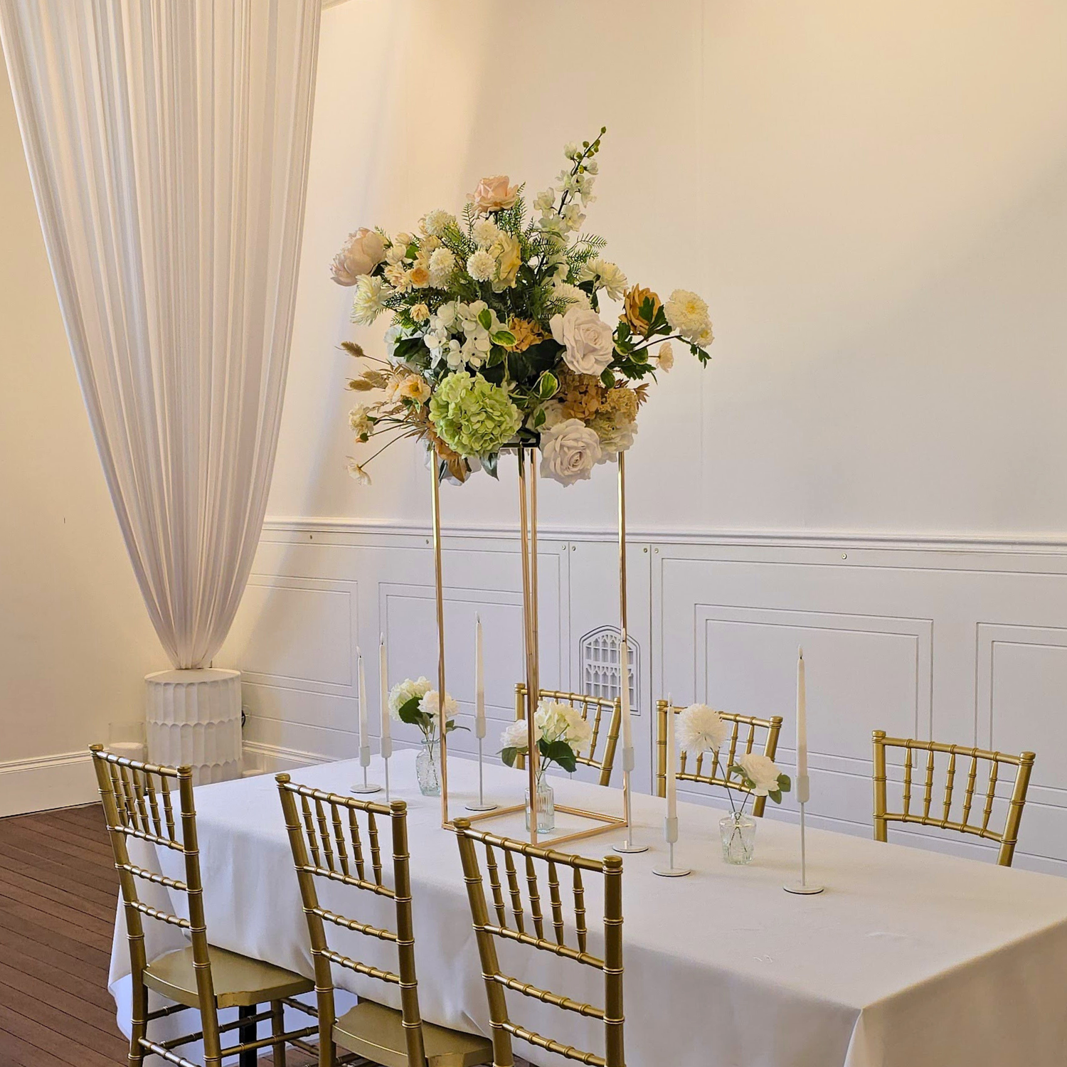 Tall floral plinth on a table with white tablecloth and gold chairs in a room with white walls and curtains. brisbane wedding guest table styling, brisbane wedding centrepieces, taper candle wedding styling brisbane, candelabra wedding centrepieces brisbane, bud vase wedding styling brisbane, tall floral plinth centrepieces brisbane, luxury guest table styling brisbane, wedding table centrepiece hire brisbane, modern wedding table styling brisbane, elegant reception table styling brisbane, wedding candles.
