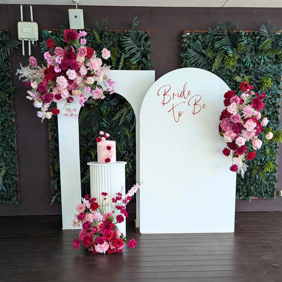 Decorative setup with floral arrangements and 'Bride to Be' sign. bridal shower backdrop, red and pink floral backdrop, elegant event styling, wedding backdrop hire, engagement party decor, Brisbane event hire, floral arch setup, romantic party backdrop, custom event decor, luxe bridal shower decorationsagainst a dark wall.