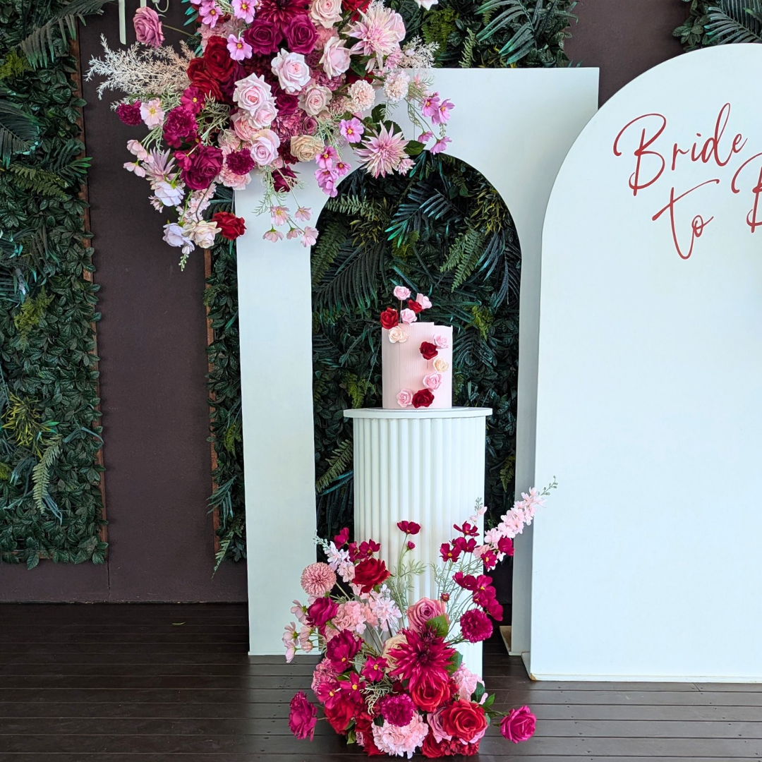 Decorative floral arrangement with a cake and 'Bride to Be' sign on a white arch. bridal shower backdrop, red and pink floral backdrop, elegant event styling, wedding backdrop hire, engagement party decor, Brisbane event hire, floral arch setup, romantic party backdrop, custom event decor, luxe bridal shower decorations