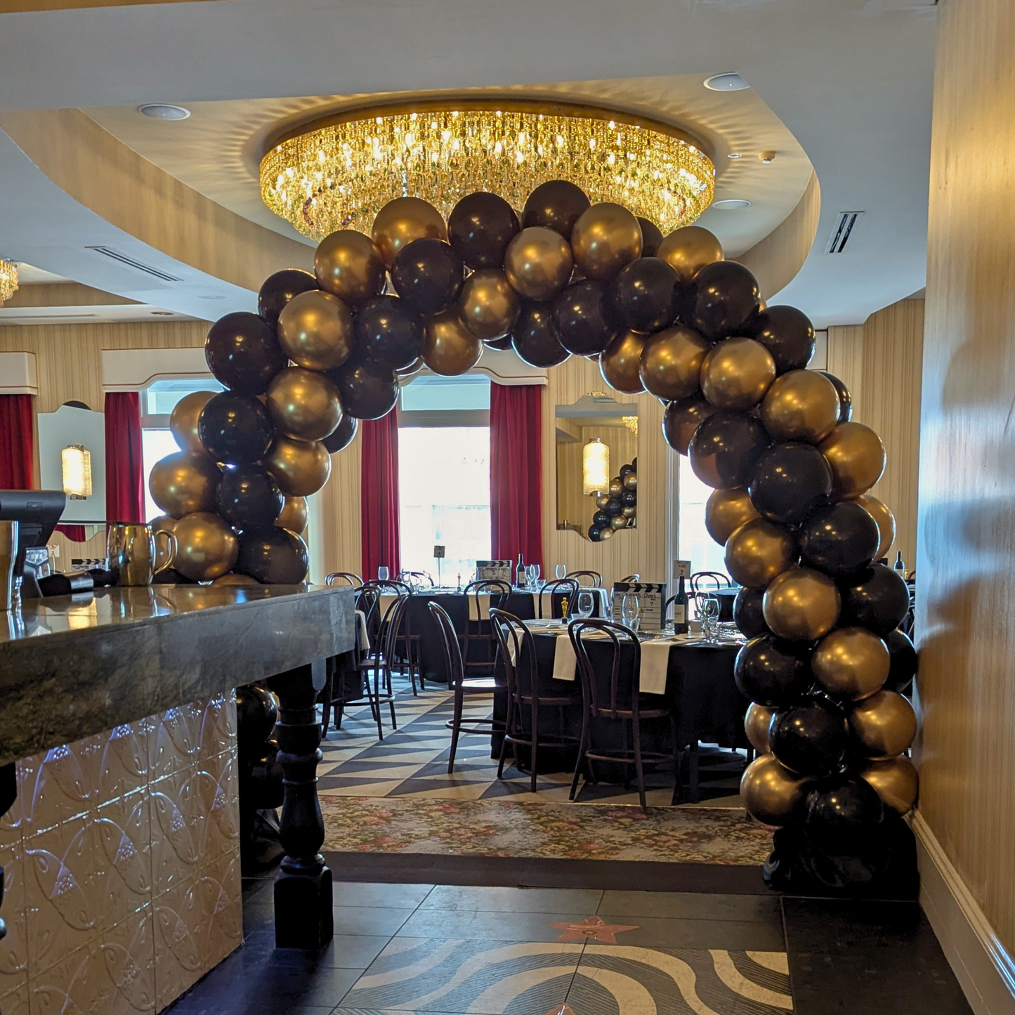 Decorative arch made of gold and black balloons in a restaurant setting with a chandelier. corporate balloon arch Brisbane, branded balloon arch QLD, corporate event styling Brisbane, balloon arch for product launch, brand activation balloon display, media wall balloon arch Brisbane, conference balloon styling QLD, corporate event balloons Gold Coast, custom colour balloon arch Brisbane, business event balloon installation QLD