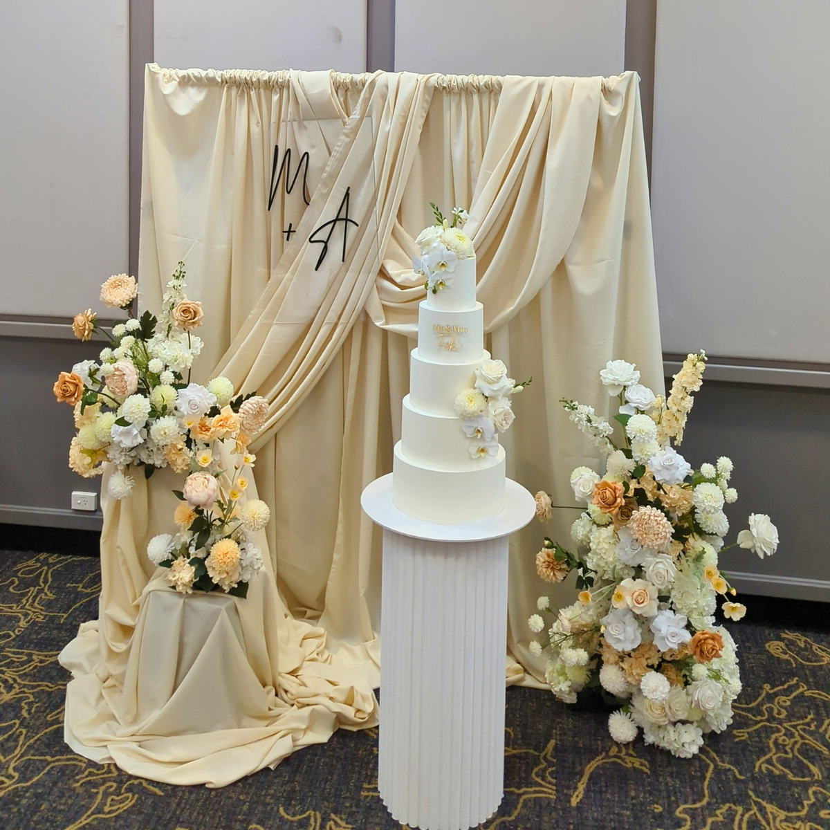 White wedding cake with floral decorations on a stand, draped fabric in the background. engagement party backdrop, draped floral backdrop, romantic event styling, Brisbane event hire, elegant engagement decor, proposal backdrop hire, floral arch setup, wedding backdrop hire, custom event backdrop, luxe engagement party decorations