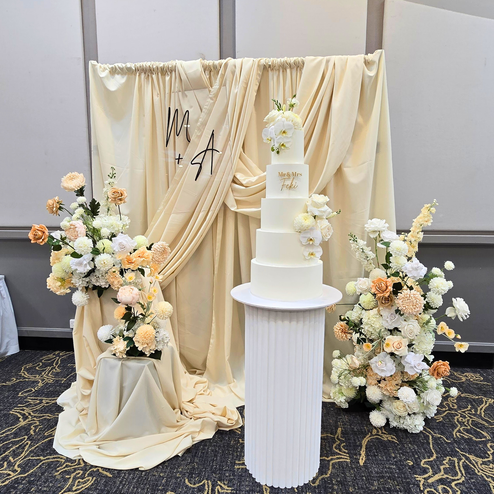 White wedding cake with floral decorations on a stand, draped fabric backdrop with 'M + A' initials. engagement party backdrop, draped floral backdrop, romantic event styling, Brisbane event hire, elegant engagement decor, proposal backdrop hire, floral arch setup, wedding backdrop hire, custom event backdrop, luxe engagement party decorations
