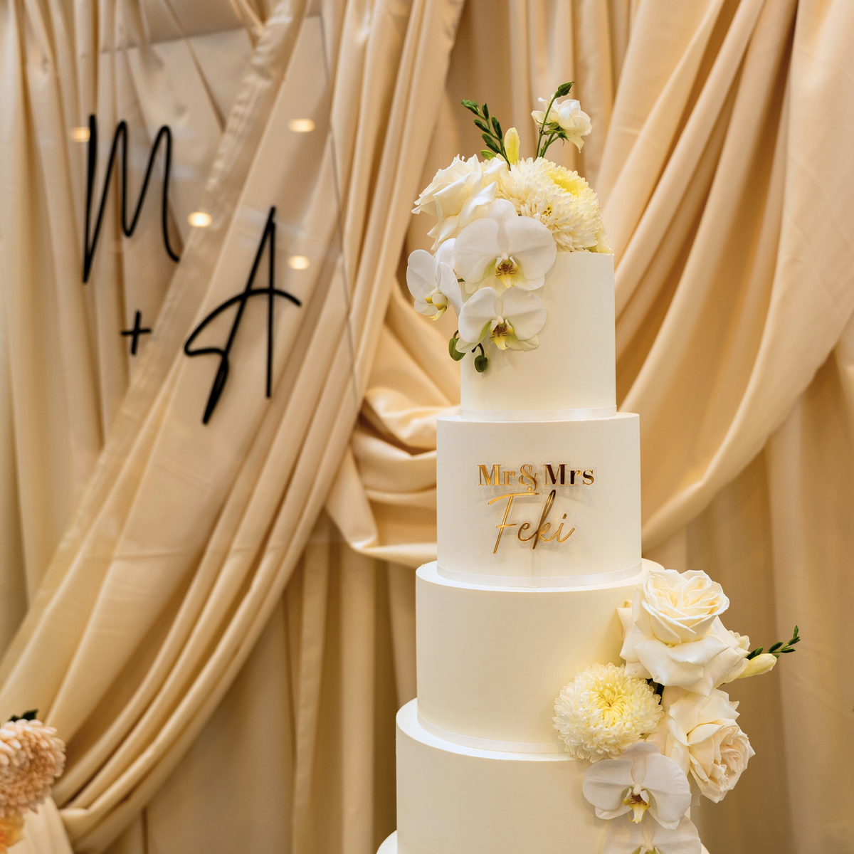 White wedding cake with floral decorations against a beige curtain backdrop. engagement party backdrop, draped floral backdrop, romantic event styling, Brisbane event hire, elegant engagement decor, proposal backdrop hire, floral arch setup, wedding backdrop hire, custom event backdrop, luxe engagement party decorations