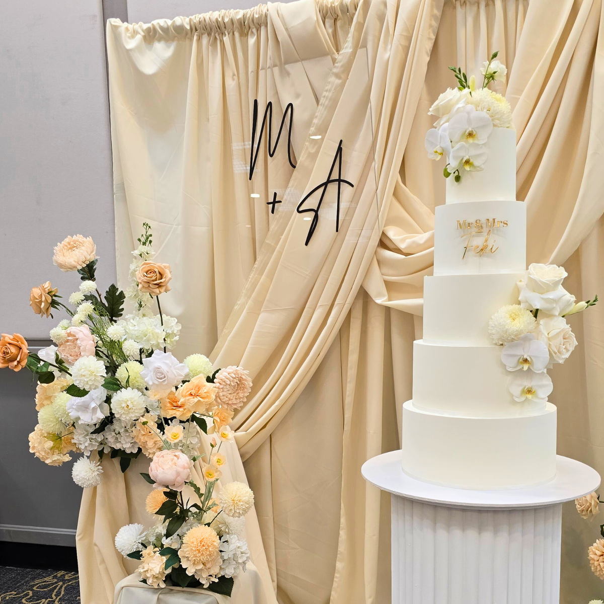 Wedding cake with floral decorations in front of a draped backdrop with 'M A' initials. engagement party backdrop, draped floral backdrop, romantic event styling, Brisbane event hire, elegant engagement decor, proposal backdrop hire, floral arch setup, wedding backdrop hire, custom event backdrop, luxe engagement party decorations