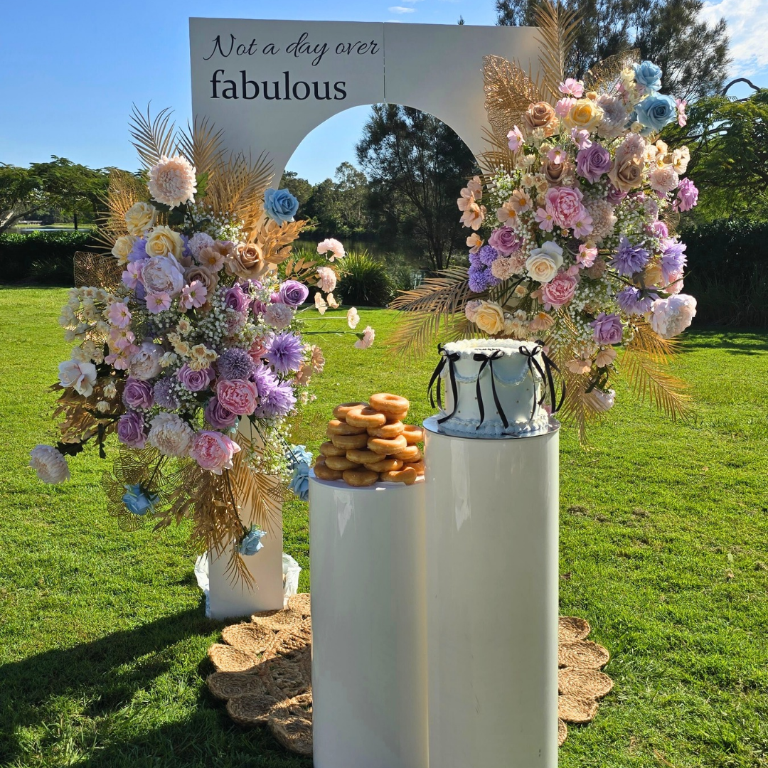 Decorative arch with flowers, cake, and donuts on a grassy outdoor setting. milestone birthday backdrop, pastel and gold floral backdrop, elegant birthday decor, Brisbane event hire, floral arch setup, luxe birthday styling, custom event backdrop, 30th birthday backdrop hire, 40th birthday decor ideas, pastel floral party setup