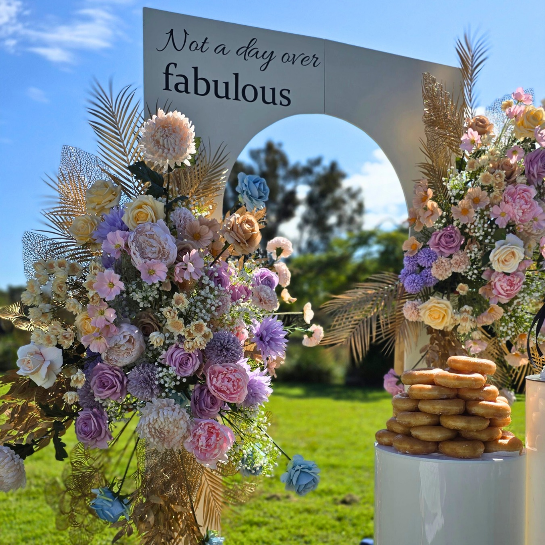 Decorative arch with flowers and 'Not a day over fabulous' sign outdoors. milestone birthday backdrop, pastel and gold floral backdrop, elegant birthday decor, Brisbane event hire, floral arch setup, luxe birthday styling, custom event backdrop, 30th birthday backdrop hire, 40th birthday decor ideas, pastel floral party setup