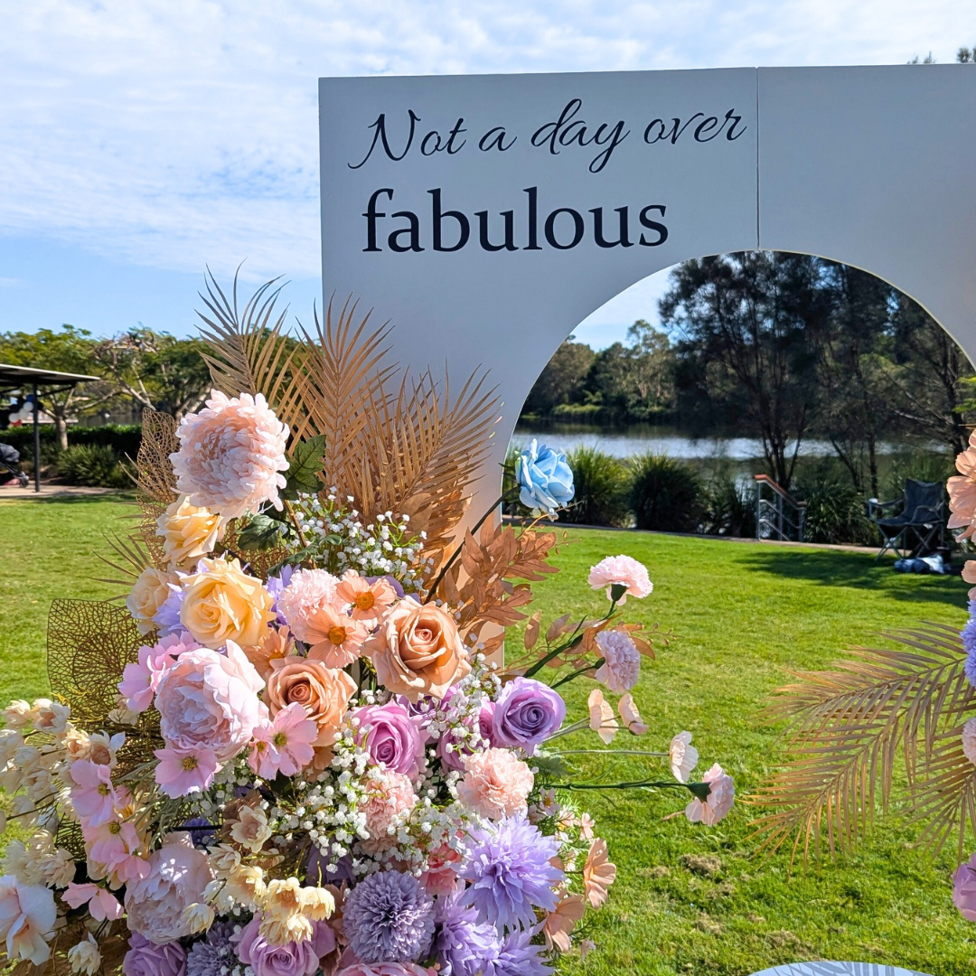Floral arrangement with 'Not a day over fabulous' sign in an outdoor setting. milestone birthday backdrop, pastel and gold floral backdrop, elegant birthday decor, Brisbane event hire, floral arch setup, luxe birthday styling, custom event backdrop, 30th birthday backdrop hire, 40th birthday decor ideas, pastel floral party setup