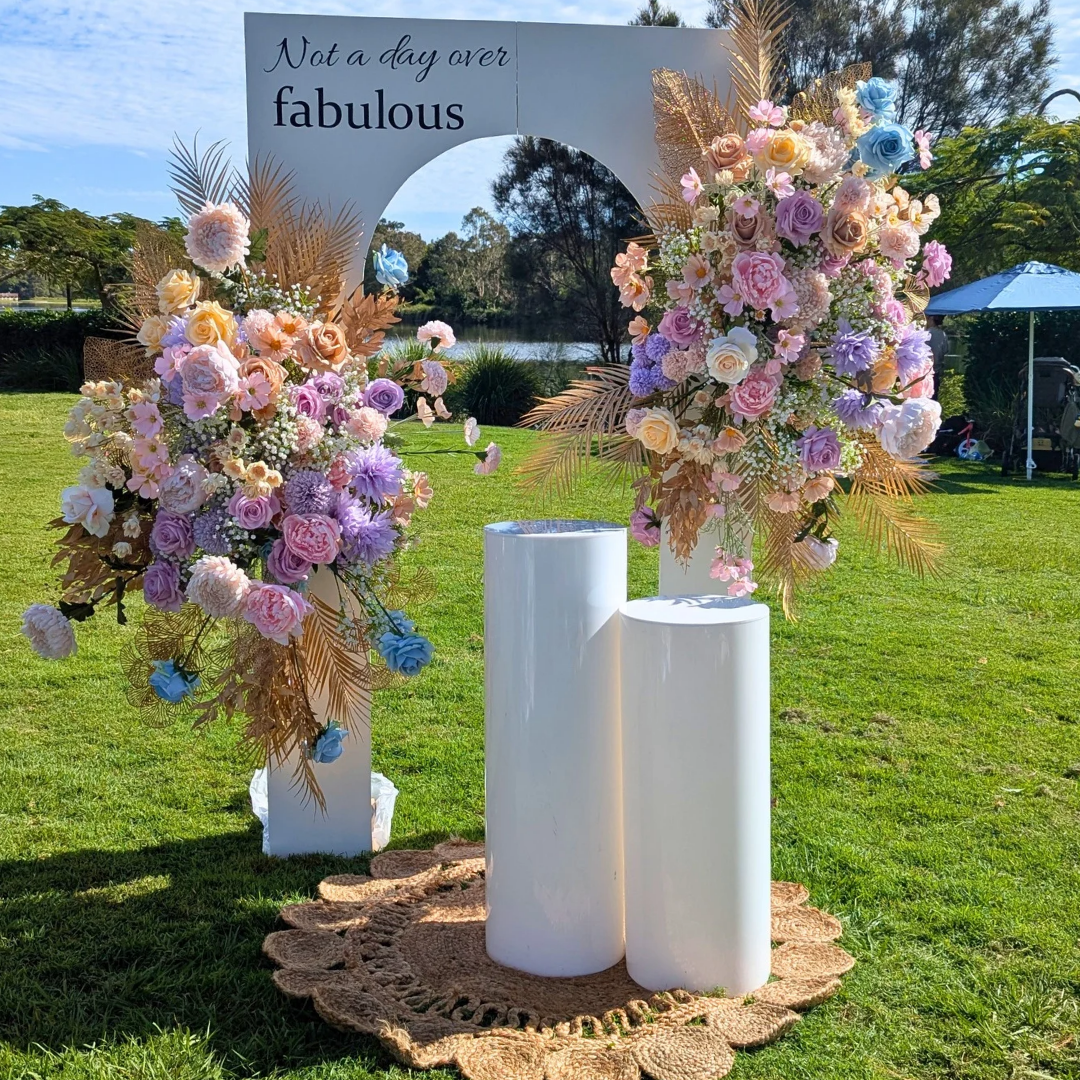 Floral arch with pastel flowers and white columns on a grassy area with a sign in the background. milestone birthday backdrop, pastel and gold floral backdrop, elegant birthday decor, Brisbane event hire, floral arch setup, luxe birthday styling, custom event backdrop, 30th birthday backdrop hire, 40th birthday decor ideas, pastel floral party setup