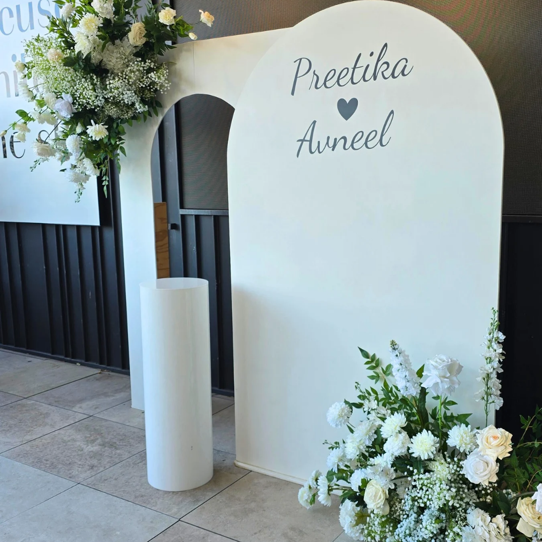 White arch with 'Preetika Avneel' text, surrounded by floral arrangements indoors. engagement party backdrop, floral engagement backdrop, romantic event styling, wedding decor hire, proposal backdrop hire, Brisbane event hire, elegant floral arch, custom event backdrop, pre-wedding decorations, luxe engagement party setup