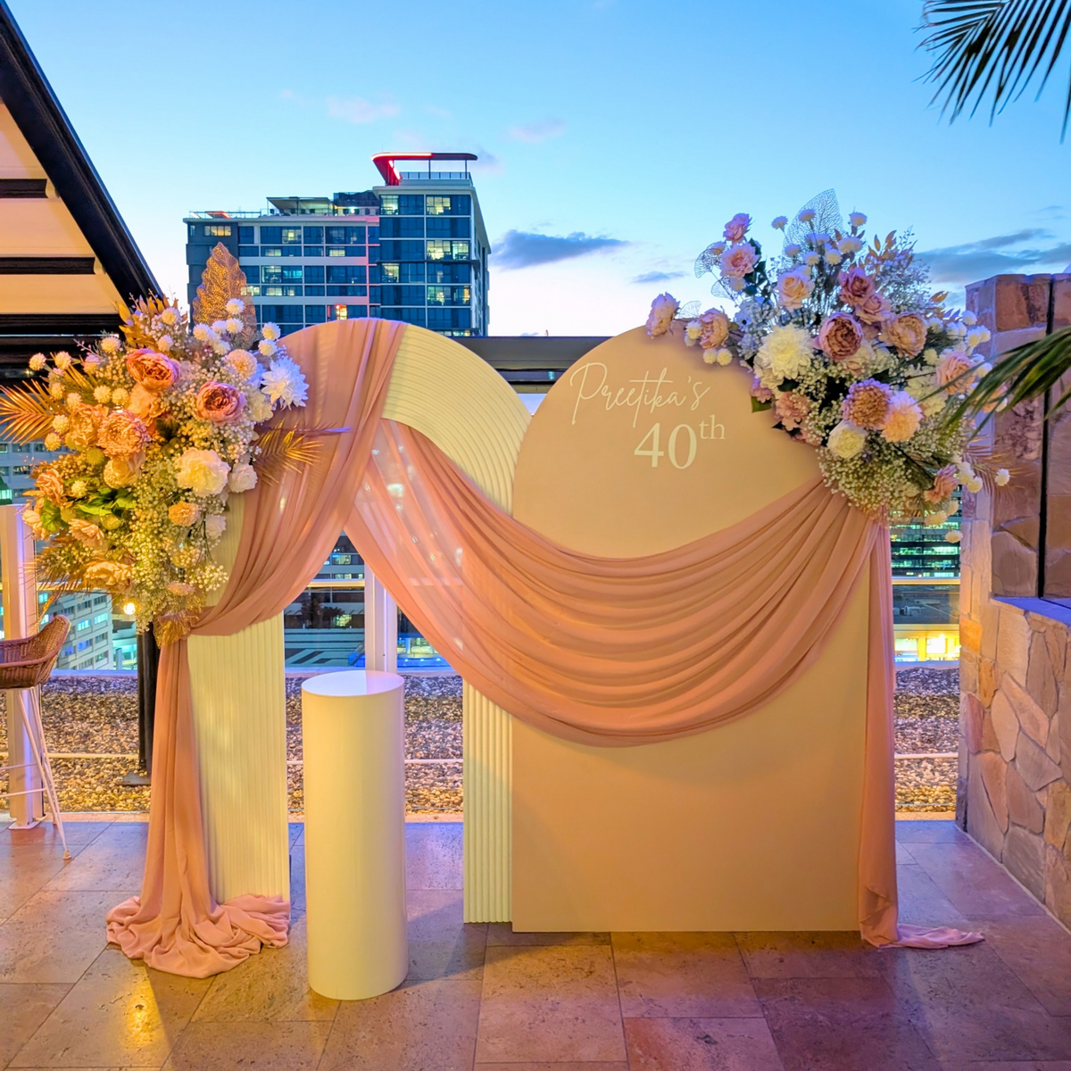 Outdoor wedding-style event backdrop with a cream arched board reading 'Preetika's 40th,' draped blush pink fabric, large floral arrangements in pinks and white, and a city skyline at dusk in the background.  Styled at Maya Rooftop bar Brisbane by Lush and Lavish Event an event styling, event planning and on-the-day coordination business. 