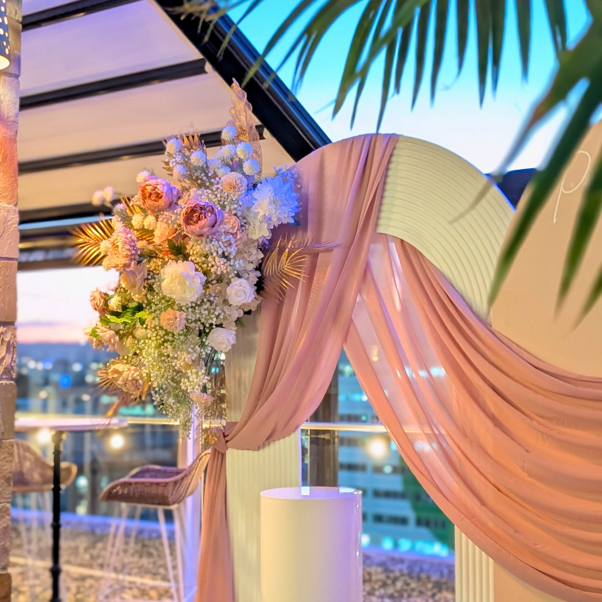 Outdoor wedding-style event backdrop with a cream arched board reading 'Preetika's 40th,' draped blush pink fabric, large floral arrangements in pinks and white, and a city skyline at dusk in the background.  Styled at Maya Rooftop bar Brisbane by Lush and Lavish Event an event styling, event planning and on-the-day coordination business. 