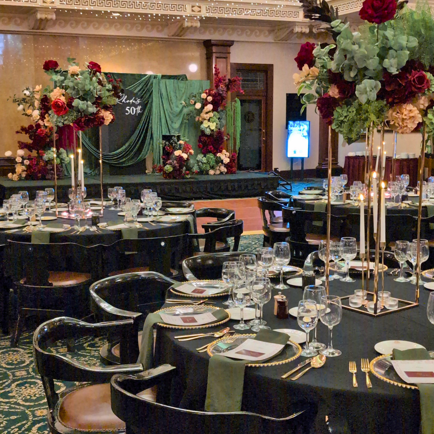 A large event hall set up for a 50th Birthday event, with round black tables covered in black tablecloths, white plates, wine glasses, and tall floral centerpieces. A green draped stage backdrop with burgundy and olive floral arrangements is visible in the background. The Tattersall Club Brisbane styled by Lush and Lavish Events.