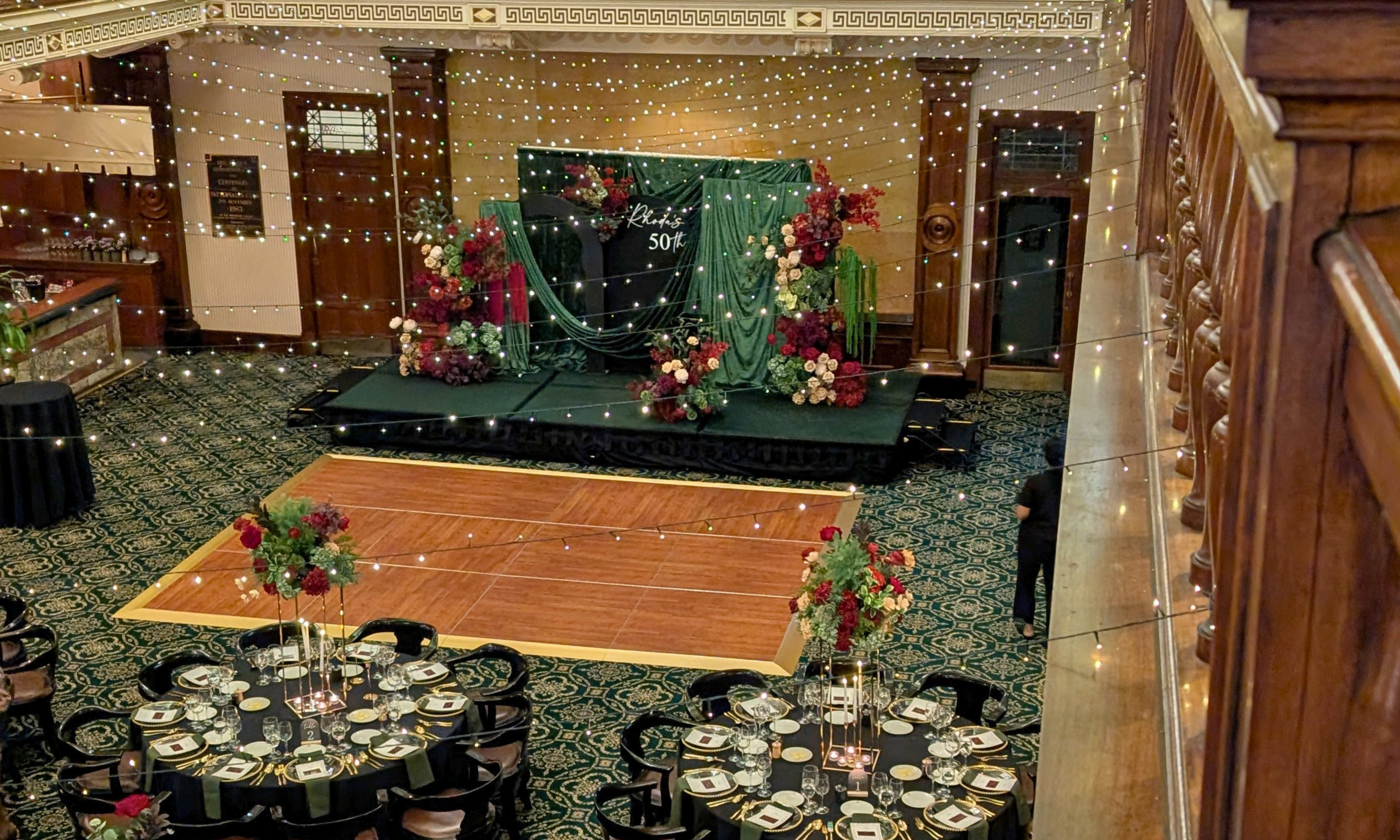 The Tattersall Club Brisbane with a decorated stage, string fairy lights, a wooden dance floor, and round dining tables set with plates and floral centerpieces. A draped backdrop with burgundy and olive floral arrangements styled by Lush and Lavish Events.