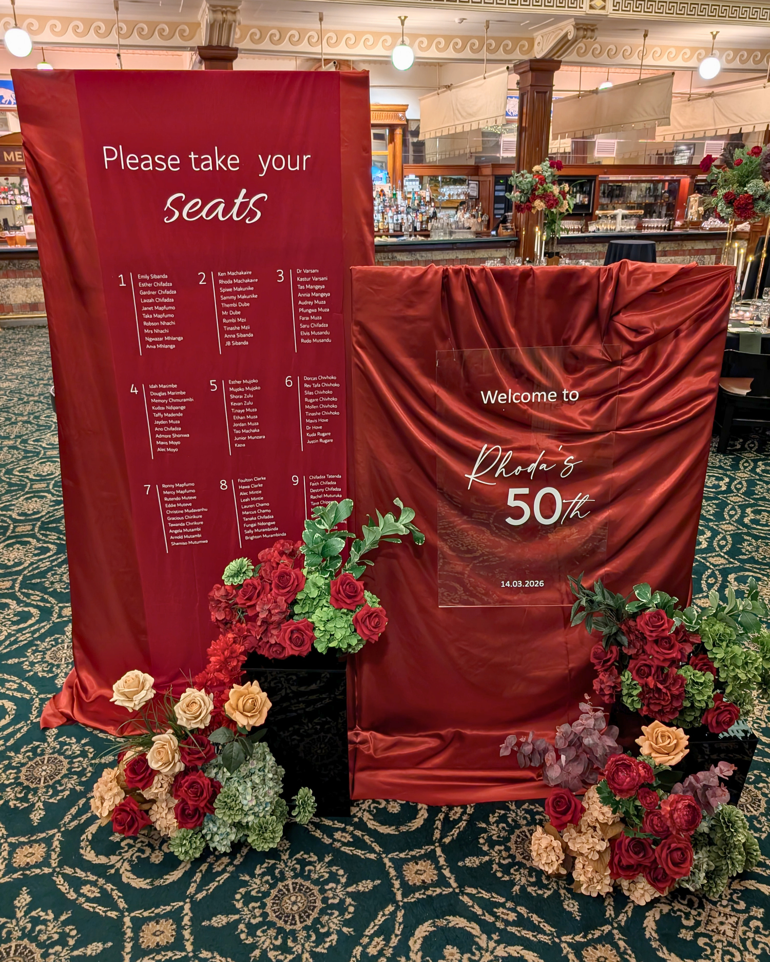 A 50th birthday celebration draped seating chart in burgundy and olive display in the Tattersall club Brisbane, featuring a large red fabric backdrop with a numbered seating list on the left and a matching red fabric welcome sign reading 'Welcome to Rhoda's 50th' on the right, decorated with red, olive, and cream floral arrangements on a green patterned carpet. Event styling by Lush and Lavish Events Brisbane based event planners, event stylist and event coordinators.
