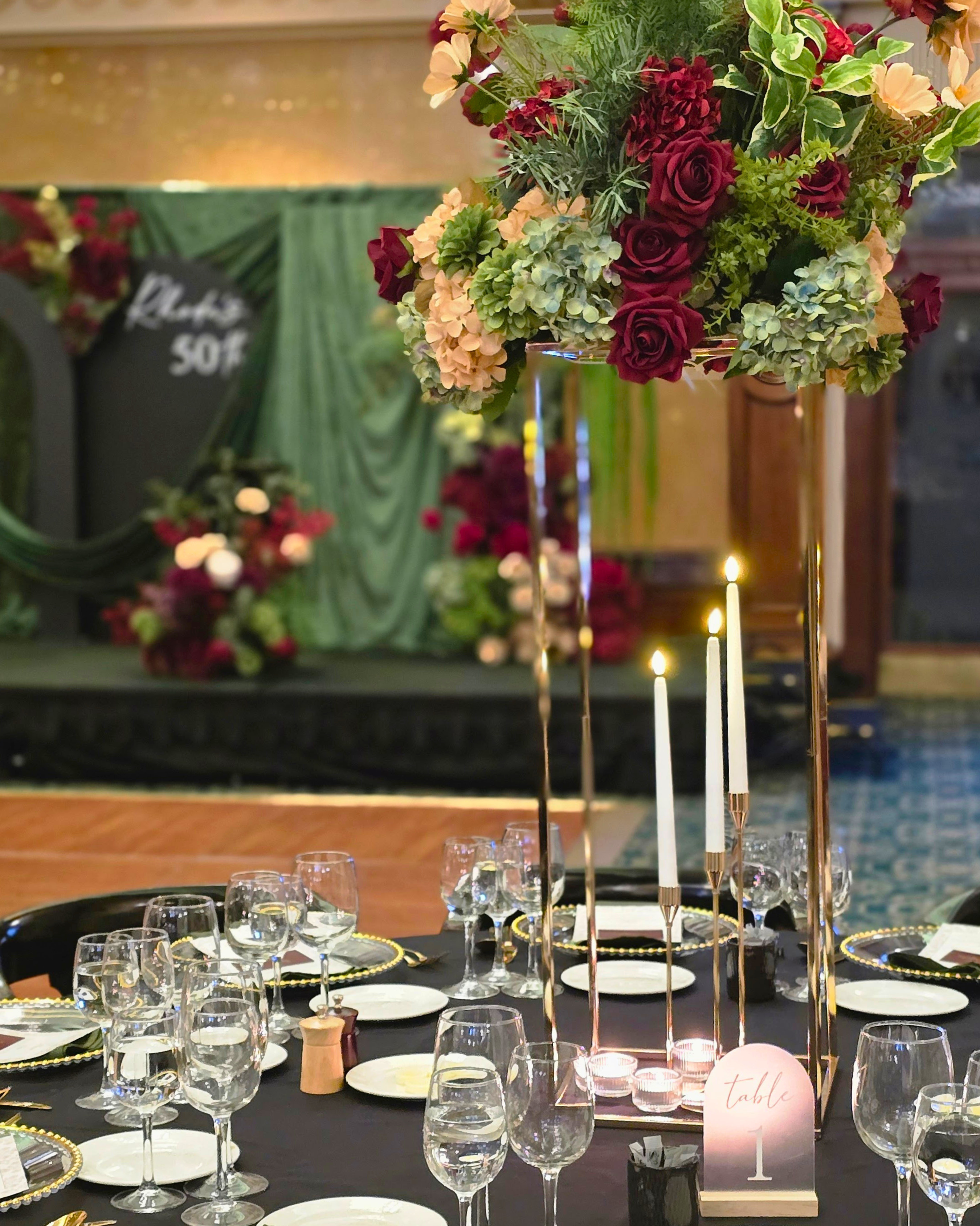 A formal event table setting with a large floral centerpiece in a tall gold stand, white plates, clear wine glasses, lit taper candles, and a table number card. Styled in The Tattersall Club Brisbane by Brisbane based Event stylists, event planners and event coordinators Lush and Lavish Events.