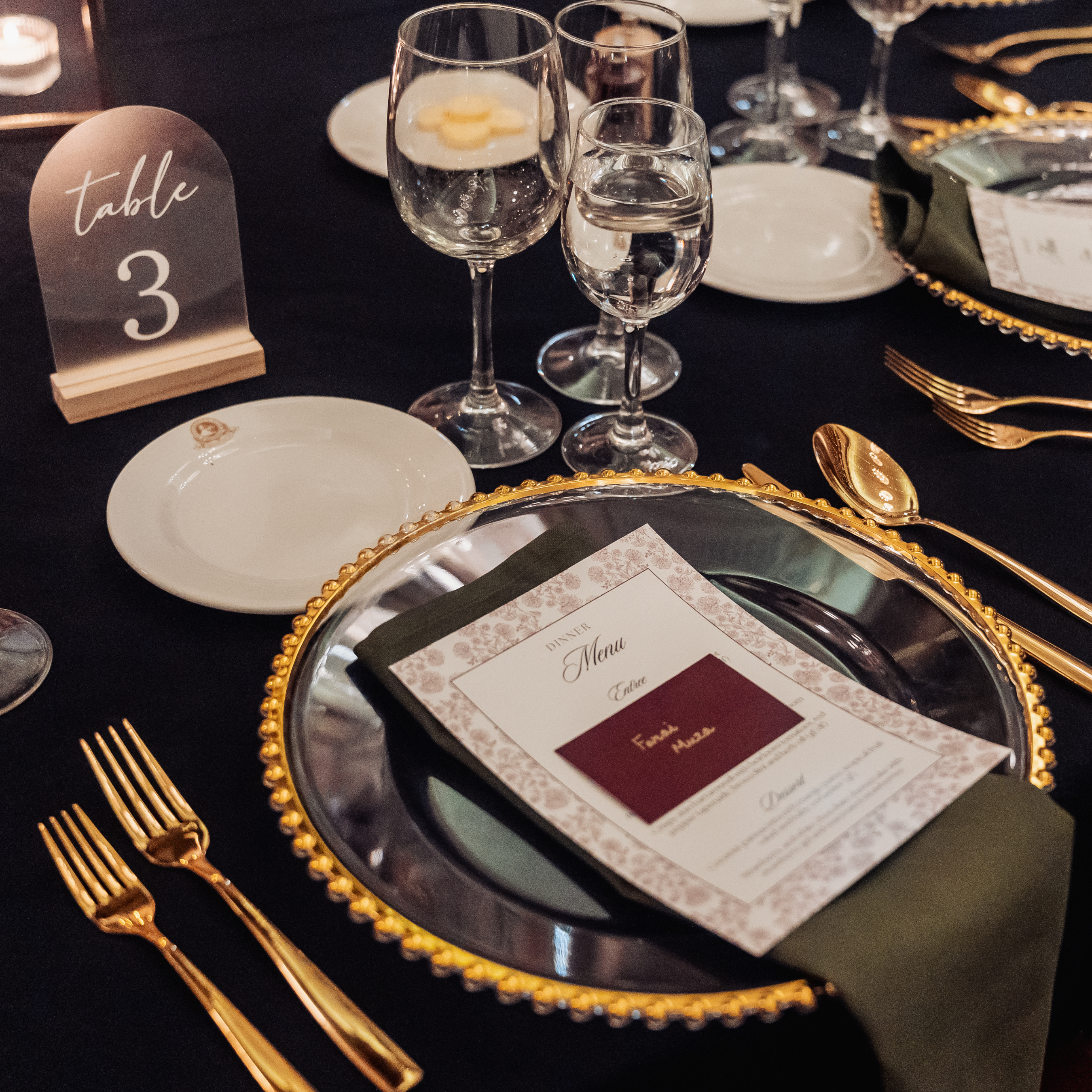 A styled milestone birthday celebration table setting with a black tablecloth, gold charger plates, gold cutlery, clear wine glasses, white plates, and a small arched table number sign reading 'table 3'. Styled in The Tattersall Club Brisbane by Brisbane based event stylists, event planners and event coordinators Lush and Lavish Events.