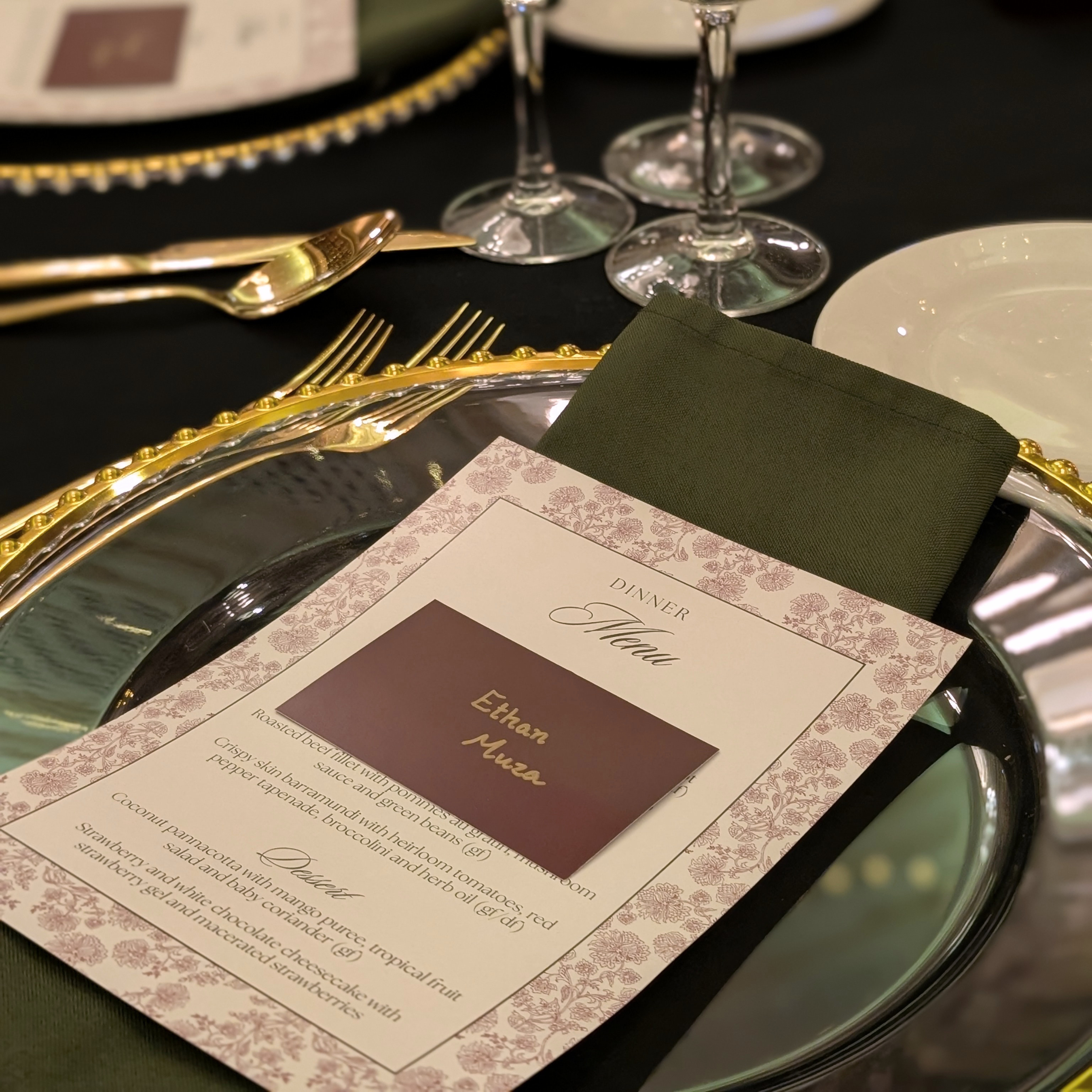 A close-up of a formal dinner place setting with a printed place card reading 'Dinner Menu Ethan Allen,' a folded dark green napkin, gold-rimmed silver charger plates, white plates, and wine glasses on a black tablecloth. Styled in The Tattersall Club Brisbane by Brisbane based Event stylists, event planners and event coordinators Lush and Lavish Events,
