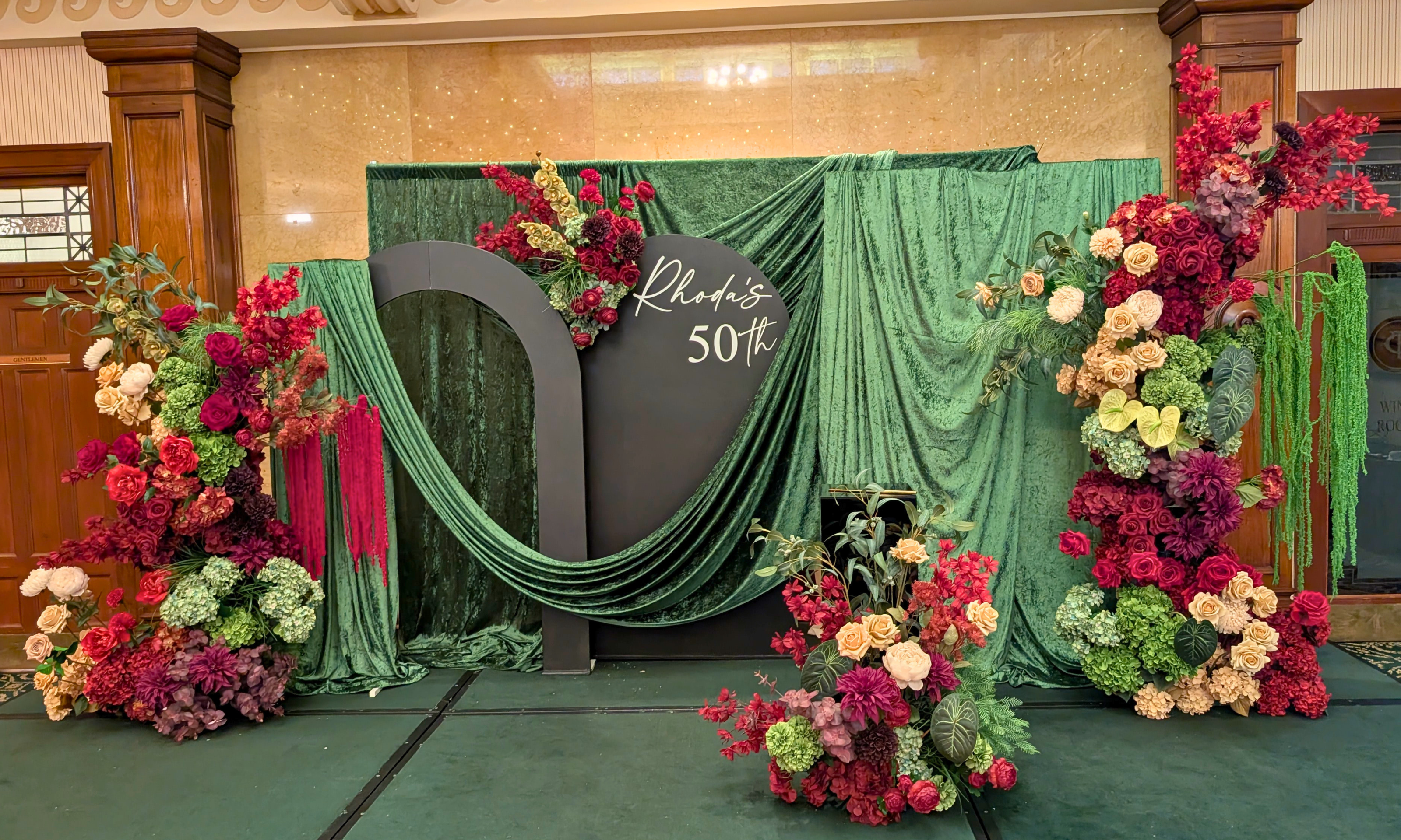 A 50th birthday event backdrop setup with a green velvet curtain, a black arch, and large floral arrangements in burgundy, cream, and olive green. The center panel reads 'Rhoda's 50th'. Styled in The Tattersal Club Brisbane by Brisbane based event stylists, event planners and event coordinators Lush and Lavish Events.