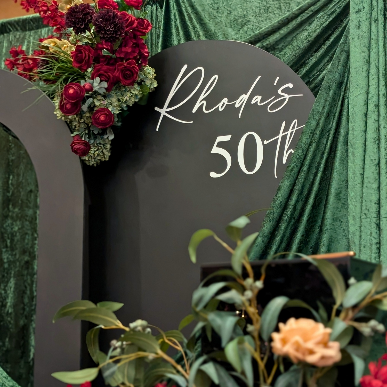 Decorative setup with flowers and 'Rhoda's 50th' sign against a green curtain. brisbane event styling, brisbane event planner, brisbane on the day coordination, brisbane 50th birthday styling, luxury event styling brisbane, brisbane birthday event planner, event stylist brisbane australia, brisbane venue styling ideas, elegant birthday styling brisbane, brisbane event design services, corporate event styling brisbane, brisbane party stylist, moody event styling brisbane, brisbane event coordination services