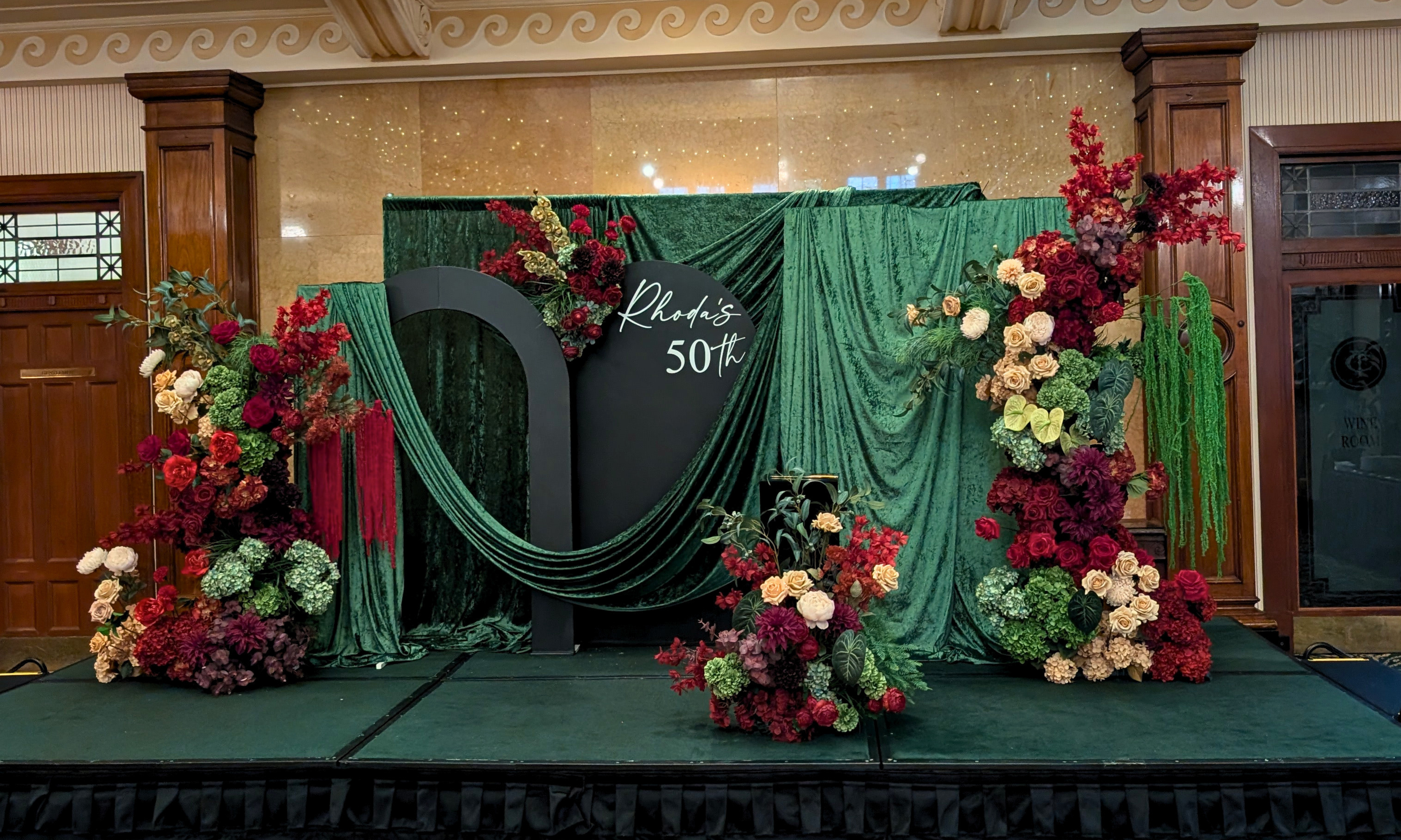 Decorative setup with floral arrangements and green drapes in a formal setting. brisbane event styling, brisbane event planner, brisbane on the day coordination, brisbane 50th birthday styling, luxury event styling brisbane, brisbane birthday event planner, event stylist brisbane australia, brisbane venue styling ideas, elegant birthday styling brisbane, brisbane event design services, corporate event styling brisbane, brisbane party stylist, moody event styling brisbane, brisbane event coordination service