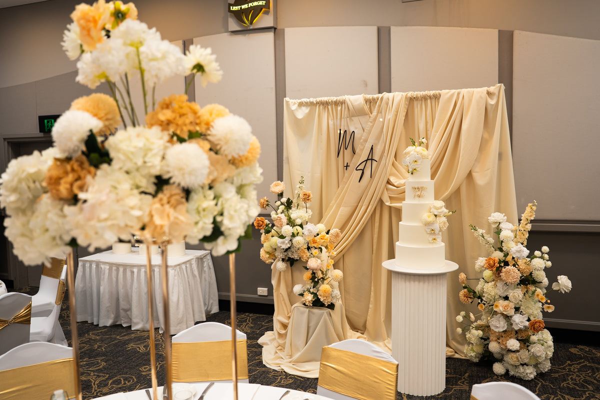 Decorative setup with flowers, a cake, and draped fabric in Kittyhawk Room, Kedron Wavell, Chermside, Brisbane. Beautiful cake table decoration with rippled plinth to hold the multi-story wedding cake. Luxurious draped backdrop for photos. 

Brisbane weddings styling, Brisbane events, Chermside events styling, Chermside wedding, Wedding reception styling. Reception Cake plinth, Wedding cake styling.